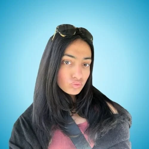 A woman with long black hair wearing sunglasses on her head, making a pouty face, against a blue background.
