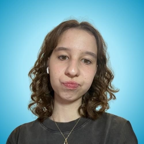 Young woman with curly brown hair, wearing a black shirt, earrings, and a necklace, standing against a blue background.