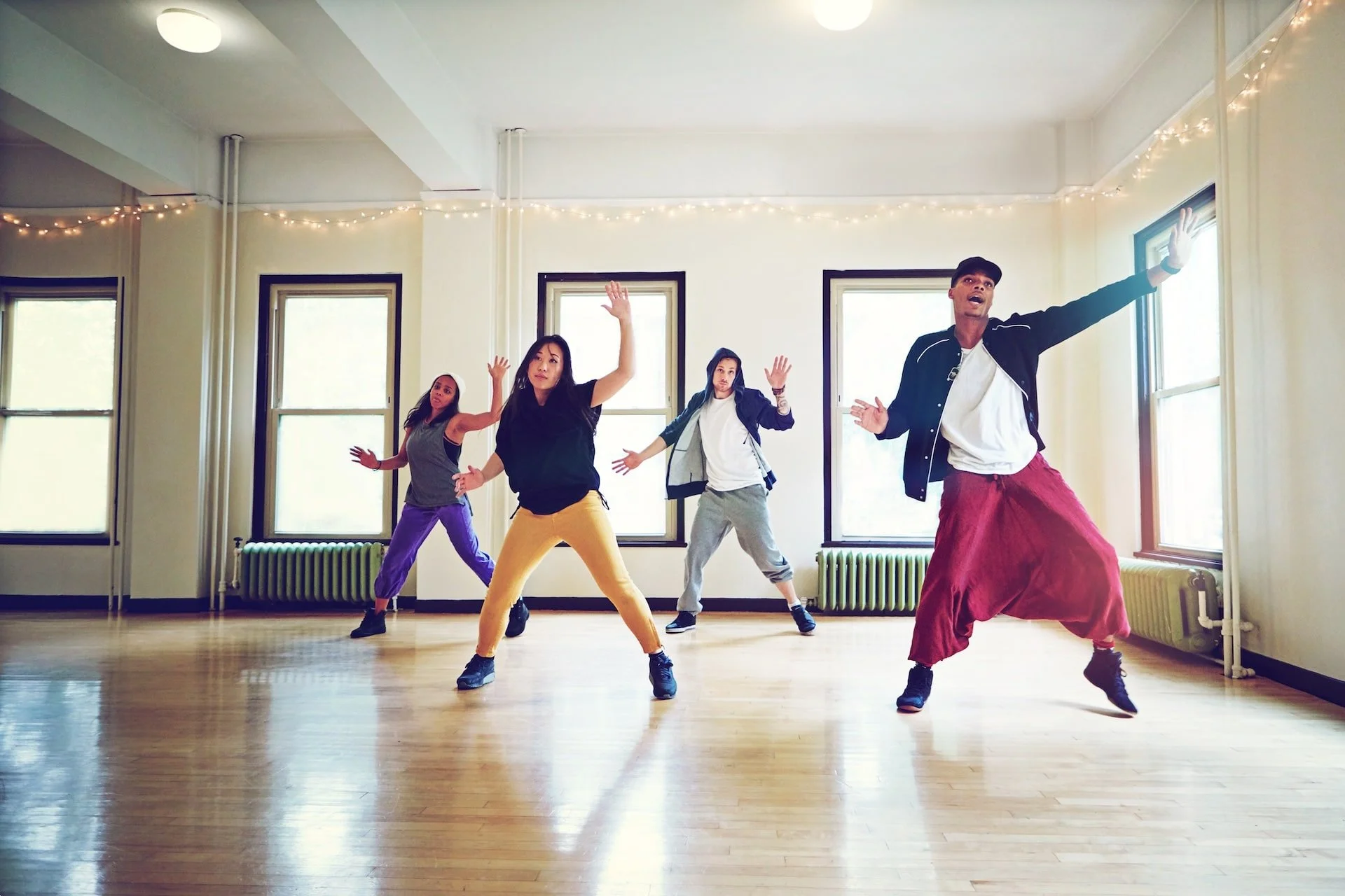 A diverse group of five people dance in a brightly lit studio with wooden floors and large windows, with fairy lights hanging from the ceiling. They are wearing casual, comfortable clothing and appear to be practicing a dance routine.