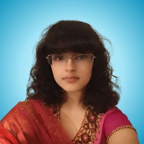 A woman with curly dark hair and glasses wearing a pink and gold traditional Indian outfit, standing against a light blue background.