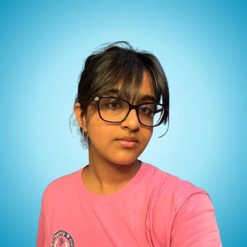 A young woman with glasses and dark hair in a messy bun, wearing a pink t-shirt with a logo, against a blue background.