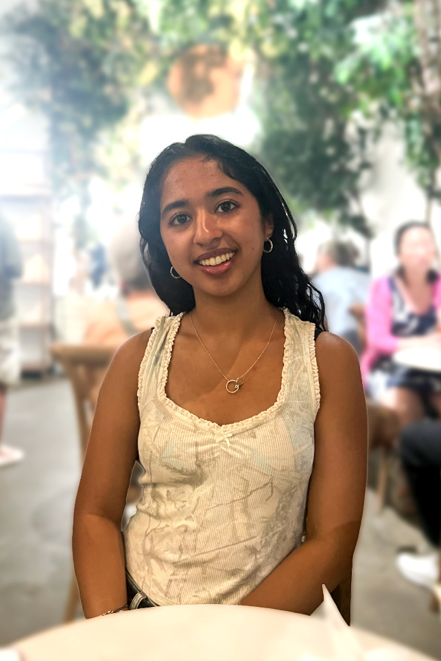 A young woman with dark hair wearing a sleeveless beige top and jewelry, smiling at the camera in a bright indoor setting with blurred background of other people and greenery.