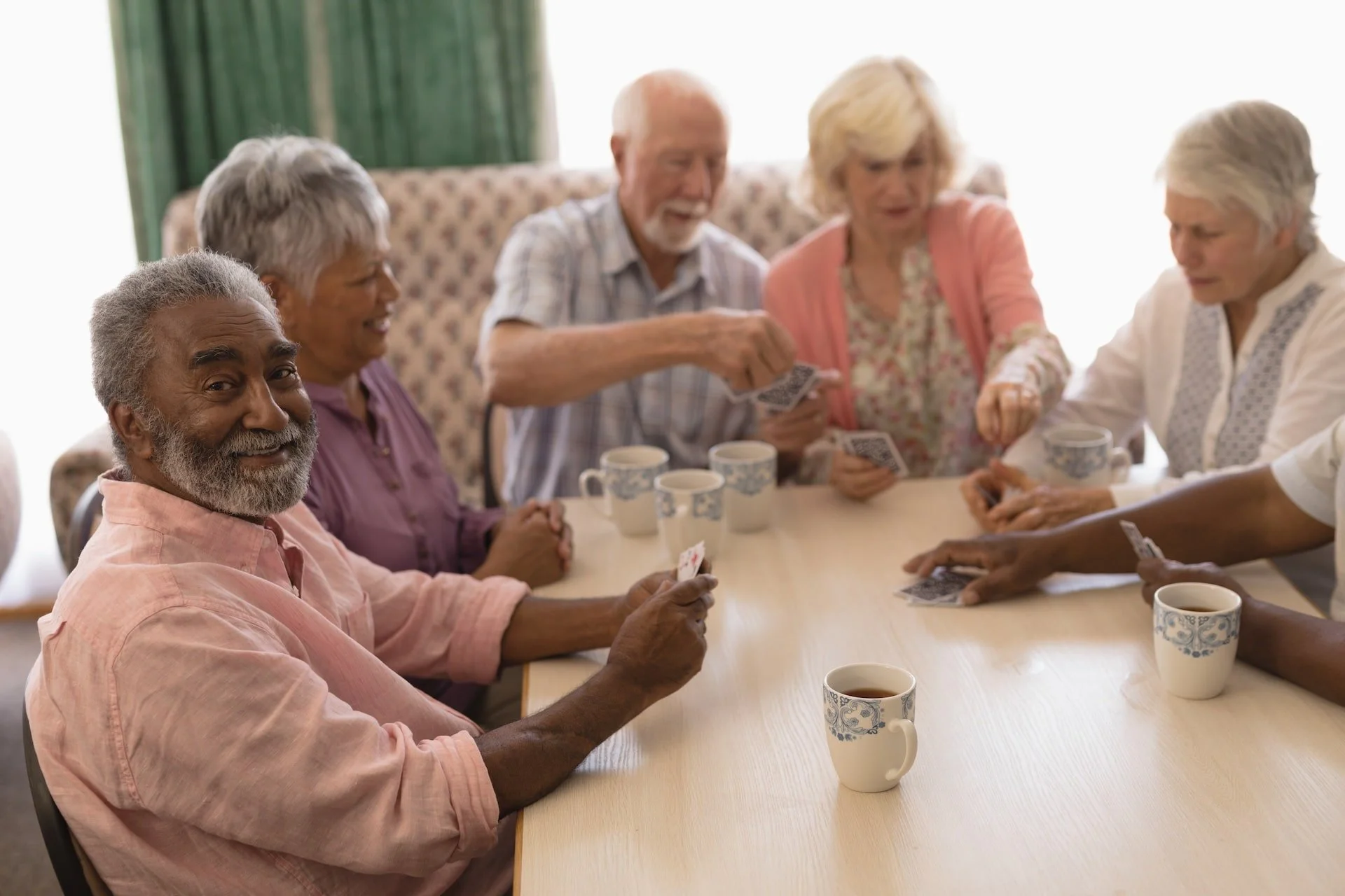 Six elderly people playing card games around a wooden table in a bright room, some smiling and holding cards, with ceramic mugs on the table.