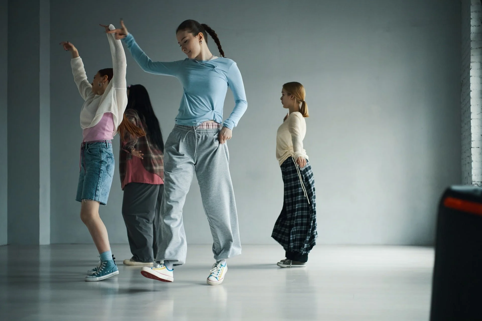 A group of young girls practicing dance moves in a spacious studio with a gray wall and wooden floor.