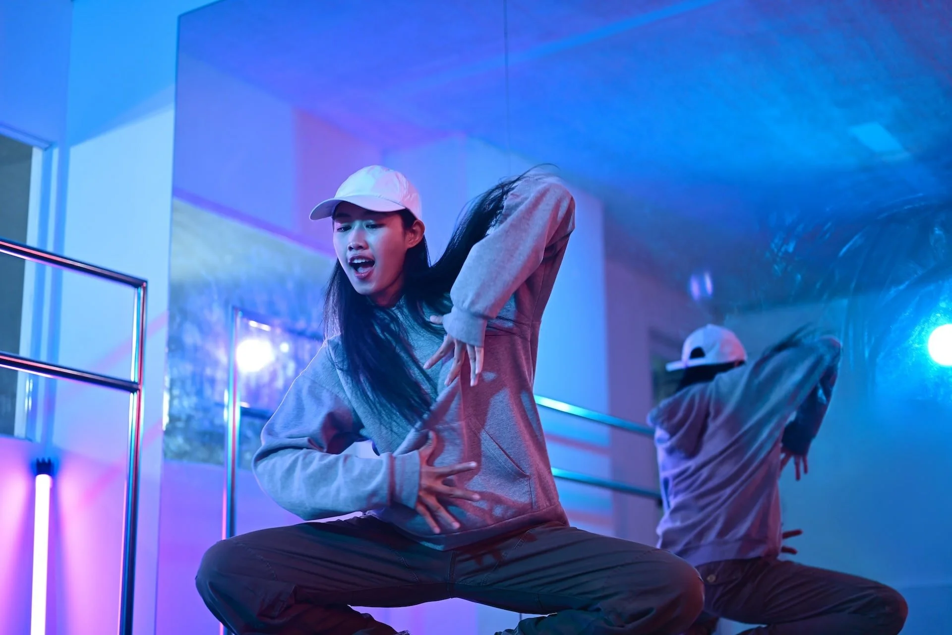 A young woman dressed in a gray hoodie and baseball cap dancing in a room with blue and purple neon lights, with another person in the background also dancing.