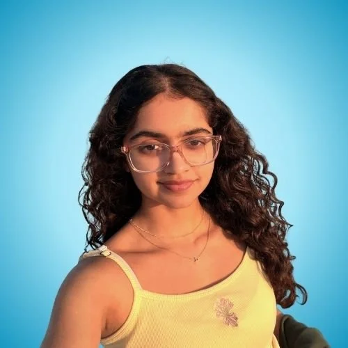A young woman with long curly brown hair and glasses, wearing a yellow tank top with a small flower design, standing against a blue background.