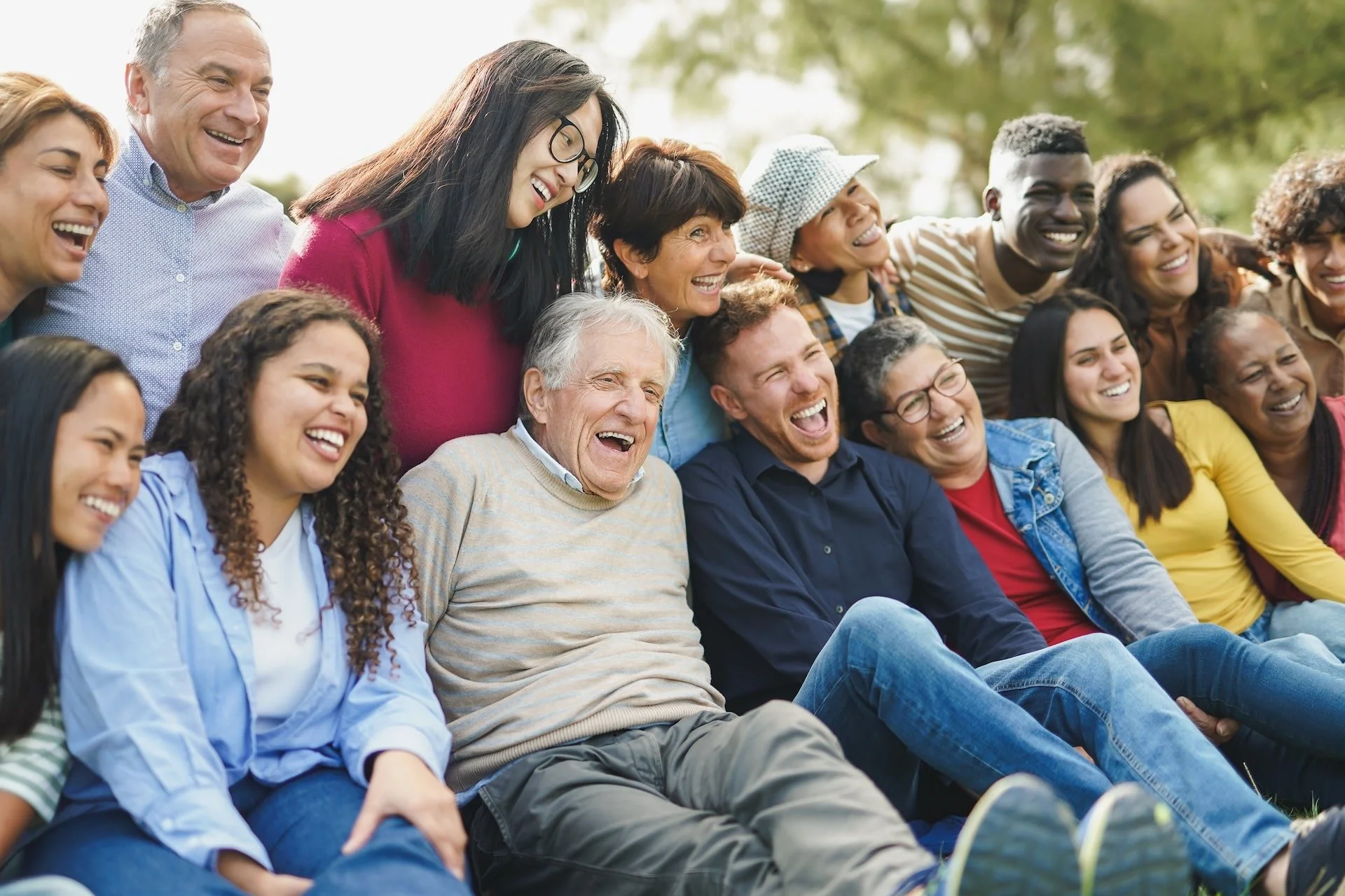 A group of diverse people outdoors, sitting together and laughing, enjoying each other's company on a sunny day.