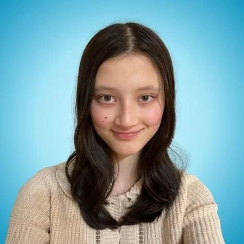 Young woman with long dark hair and fair skin smiling, wearing a beige sweater against a blue background.