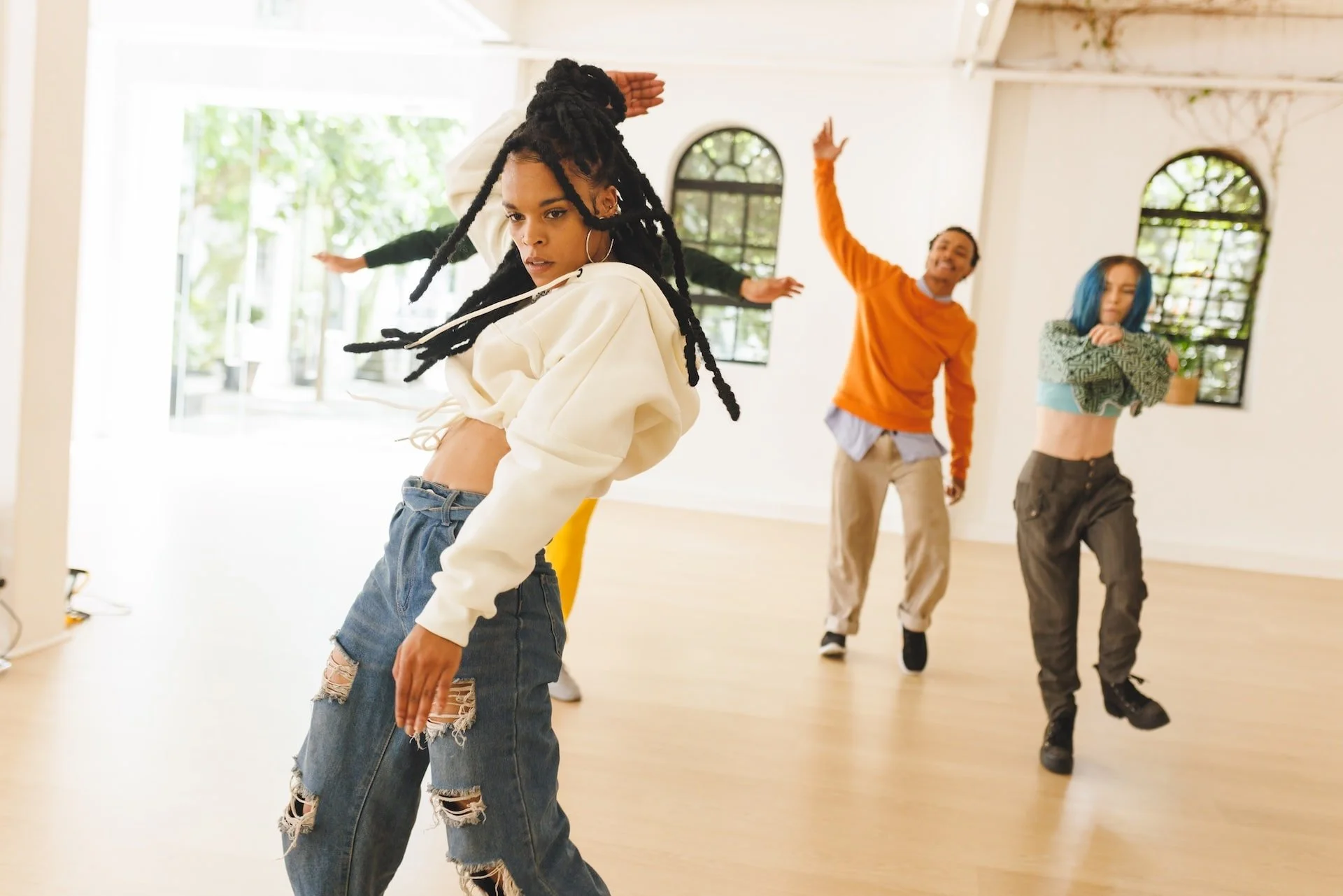 Group of diverse young dancers practicing in a bright dance studio with large windows and wood floors, displaying energetic dance moves.