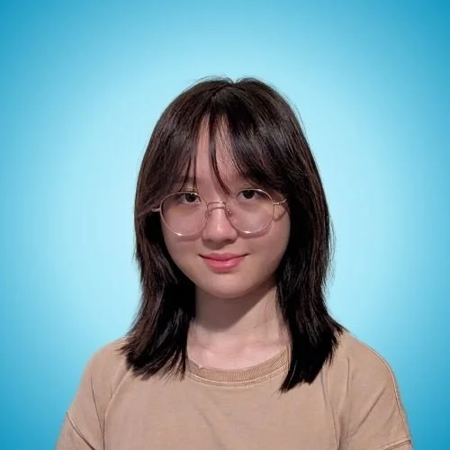 A young woman with shoulder-length dark hair, wearing round glasses and a beige t-shirt, standing against a light blue background.