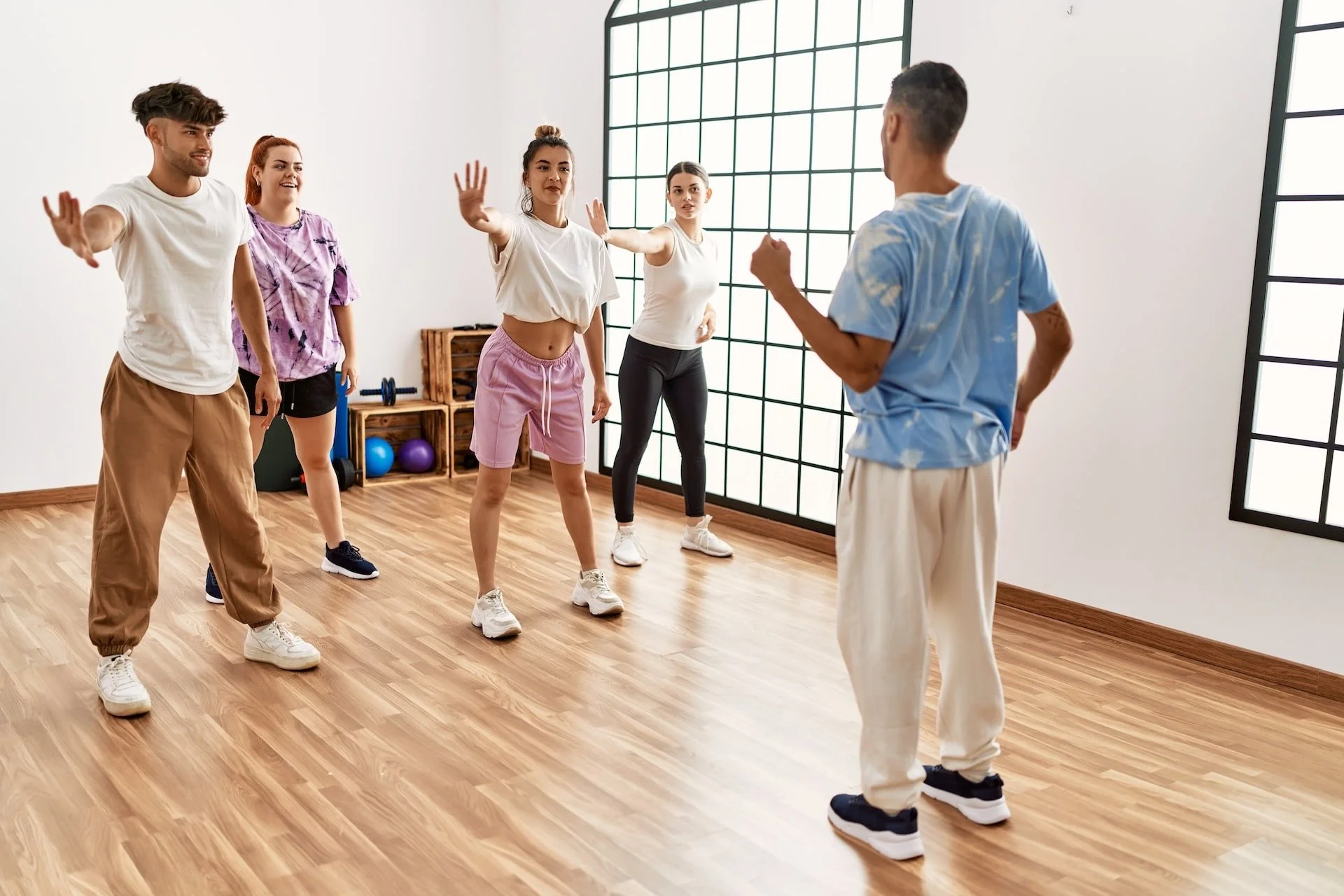 Group of five young adults participating in a dance or fitness class in a bright studio with large windows, wooden flooring, and fitness equipment in the background.