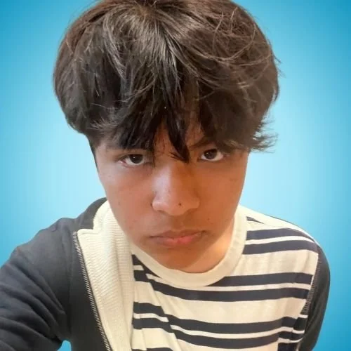 A young person with dark, messy hair and a serious expression, wearing a striped shirt and a jacket, posed against a blue background.