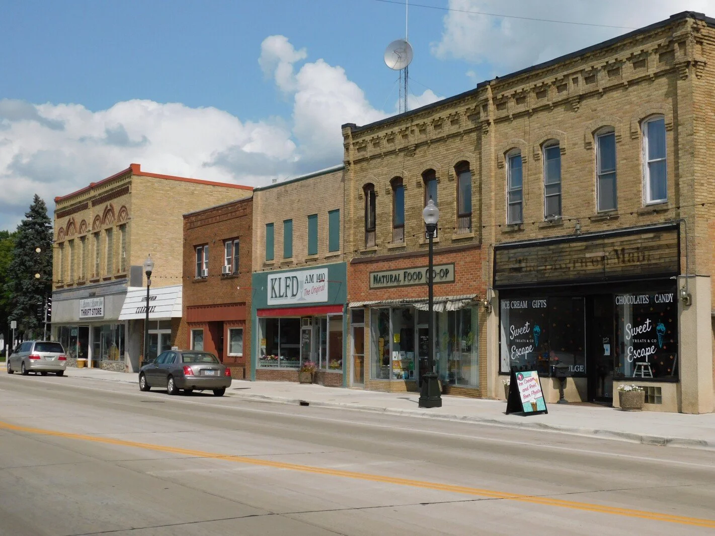 A small town main street with several historic brick storefronts, including a thrift store, a natural food shop, and an ice cream parlor, with cars parked along the curb and a partly cloudy sky overhead.
