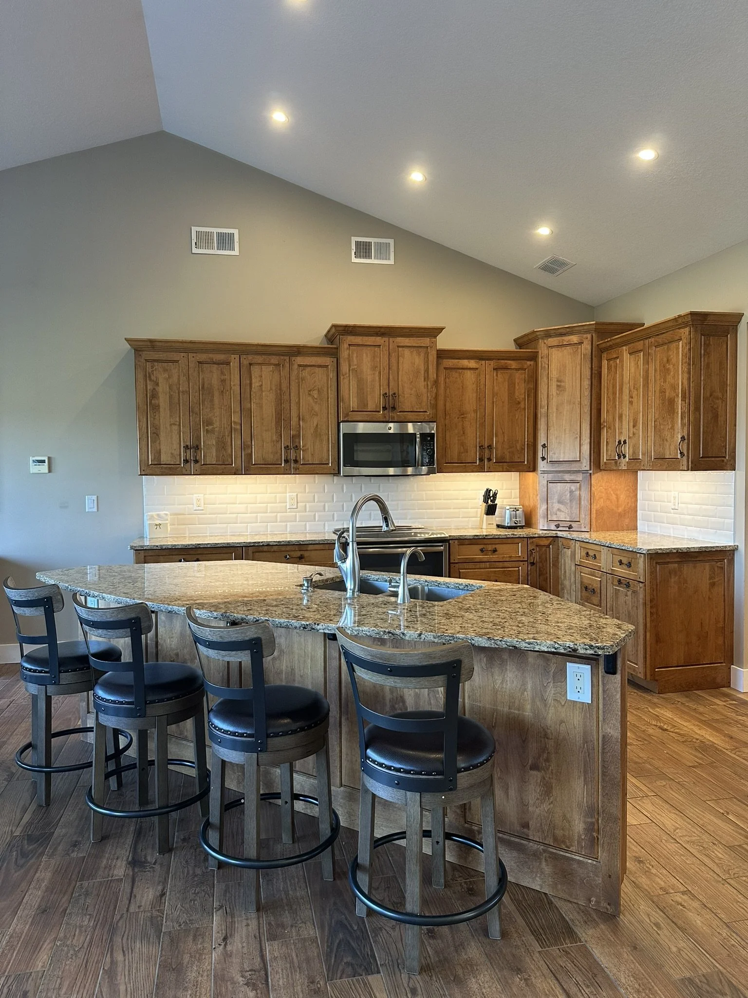 Gourmet kitchen with wooden cabinets, granite island, stainless steel appliances, and a tiled backsplash. Four black barstools are at the island.
