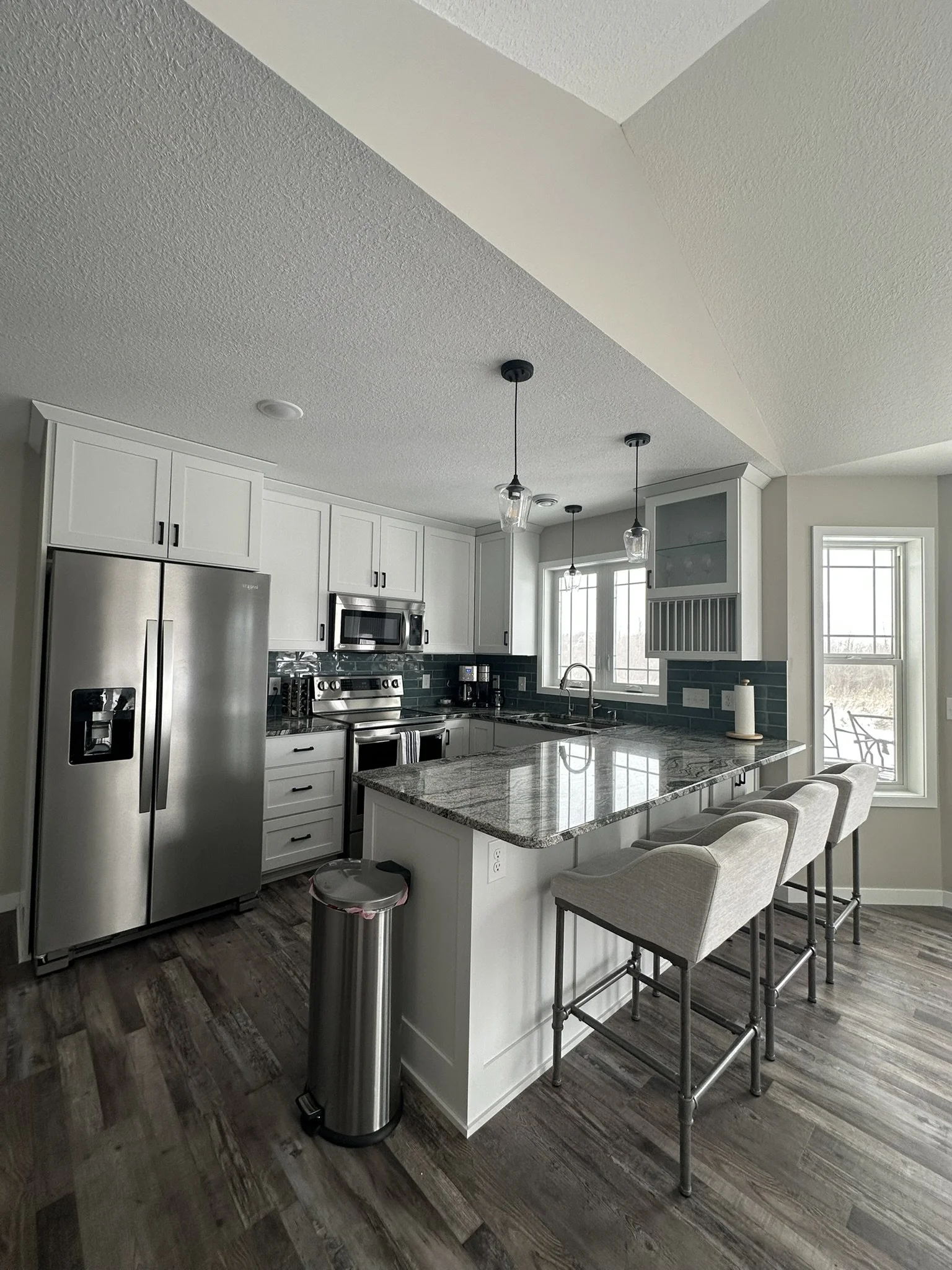 Modern kitchen with white cabinets, stainless steel appliances, granite countertop island, and beige bar stools. Natural light from large windows, wood flooring, and pendant lights hanging from the ceiling.