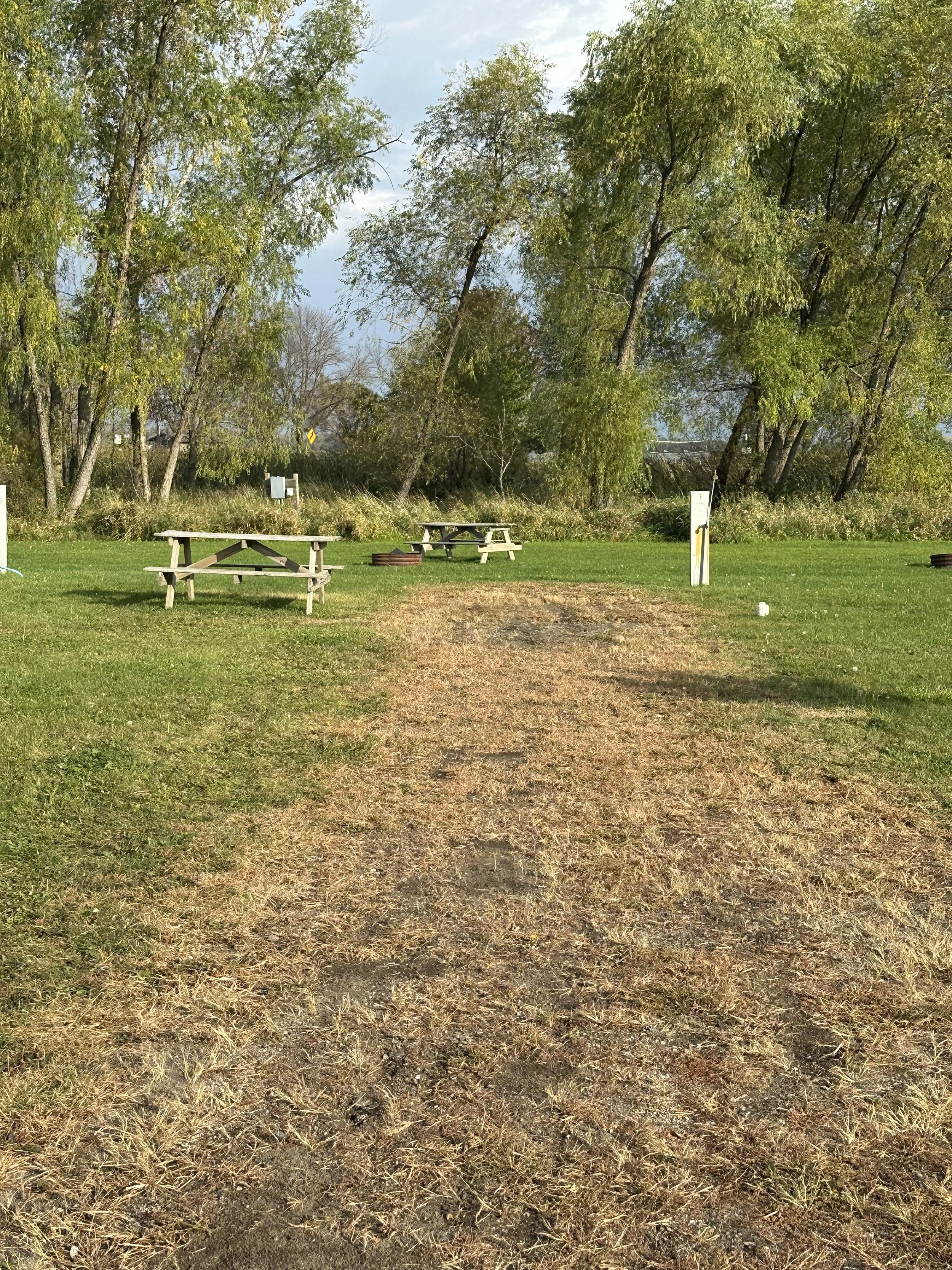 A grassy RV park area with a dirt path in the center, surrounded by trees with some leaves turning color, picnic tables, fire ring and outdoor workout equipment.