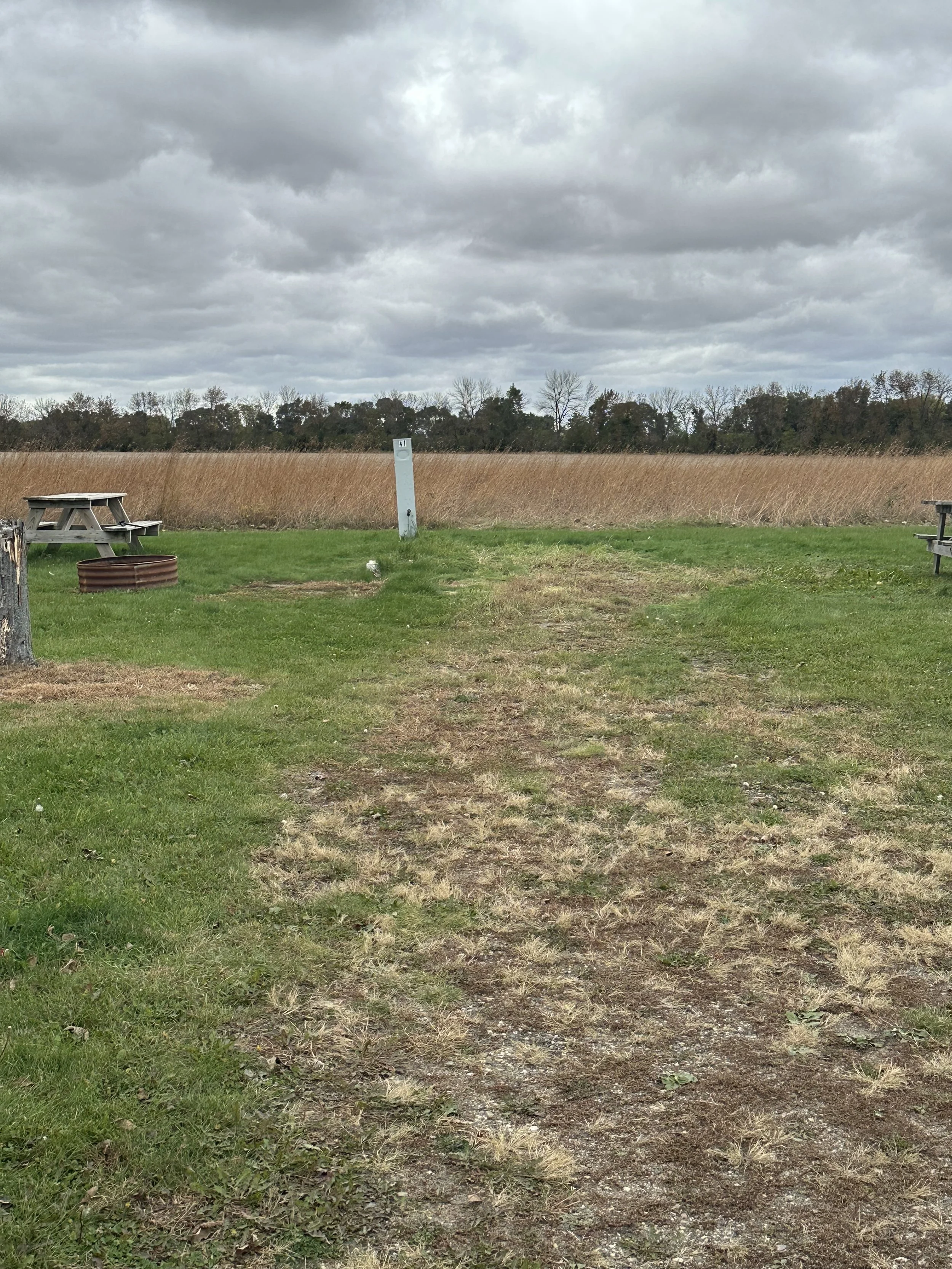 Open field under cloudy sky with patches of green and brown grass, trees in the distance, and outdoor furniture on worn grass.