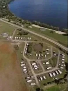 Aerial view of a shoreline RV park with a body of water, green spaces, roads, boat launch access and parked vehicles.