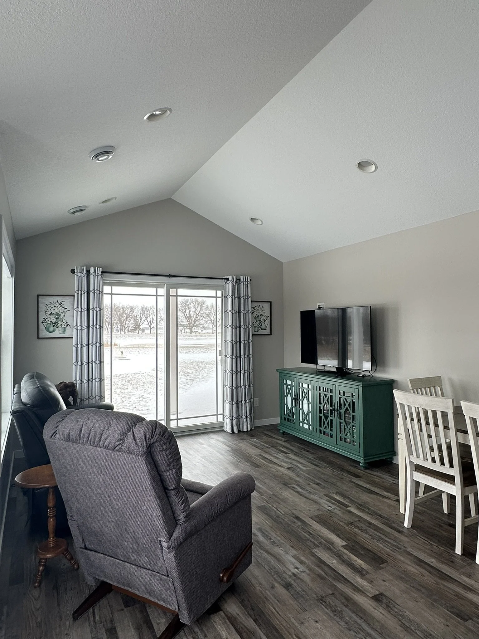 Living room with gray armchair, large window with snowy outdoor view, teal TV stand, flat-screen TV, dining table with chairs, hardwood flooring, and curtains.