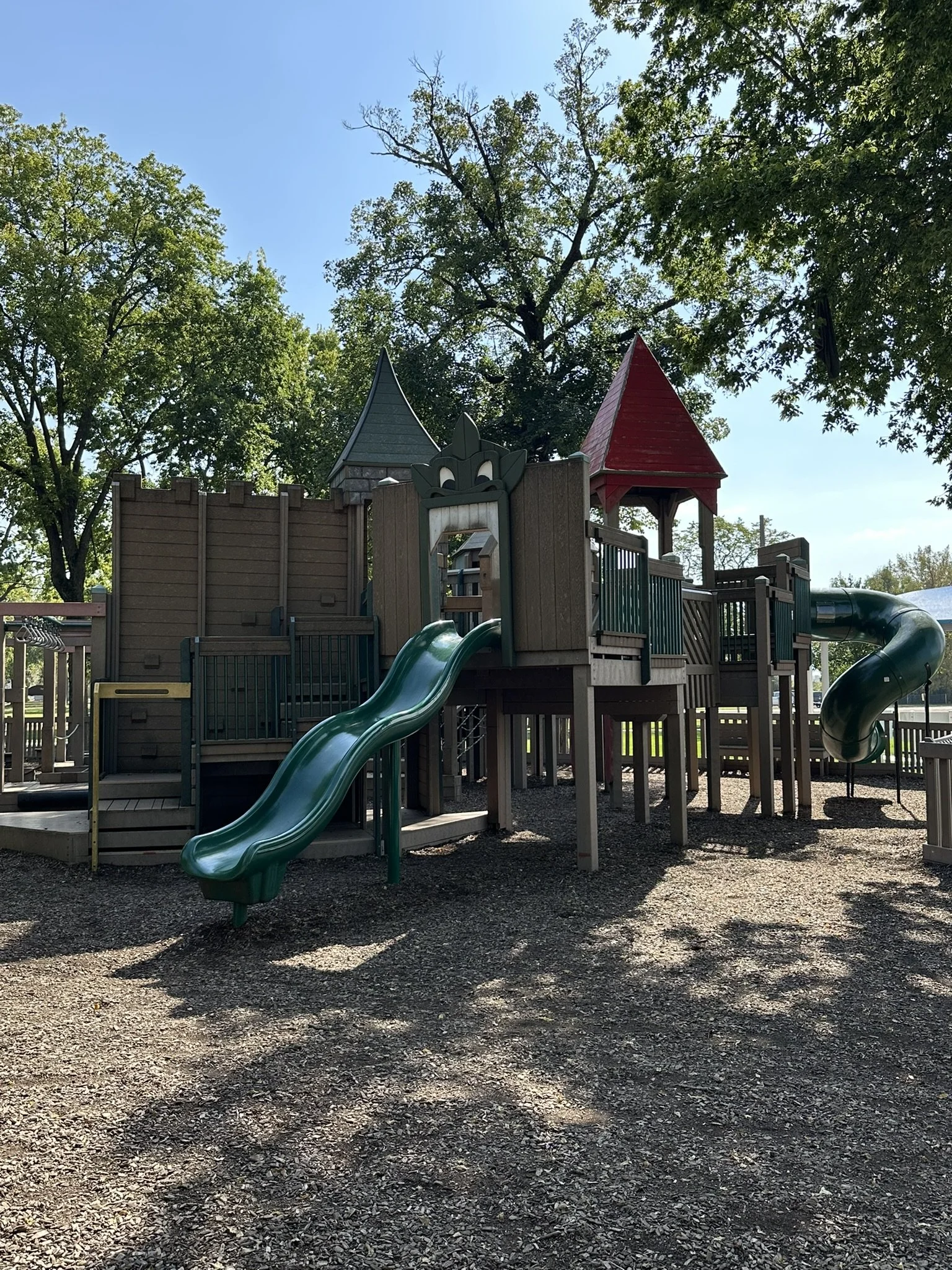 A children's playground with a wooden play structure, two slides (one green and one spiral), and two small towers with peaked roofs in gray and red. There are trees in the background and shadows on the ground.