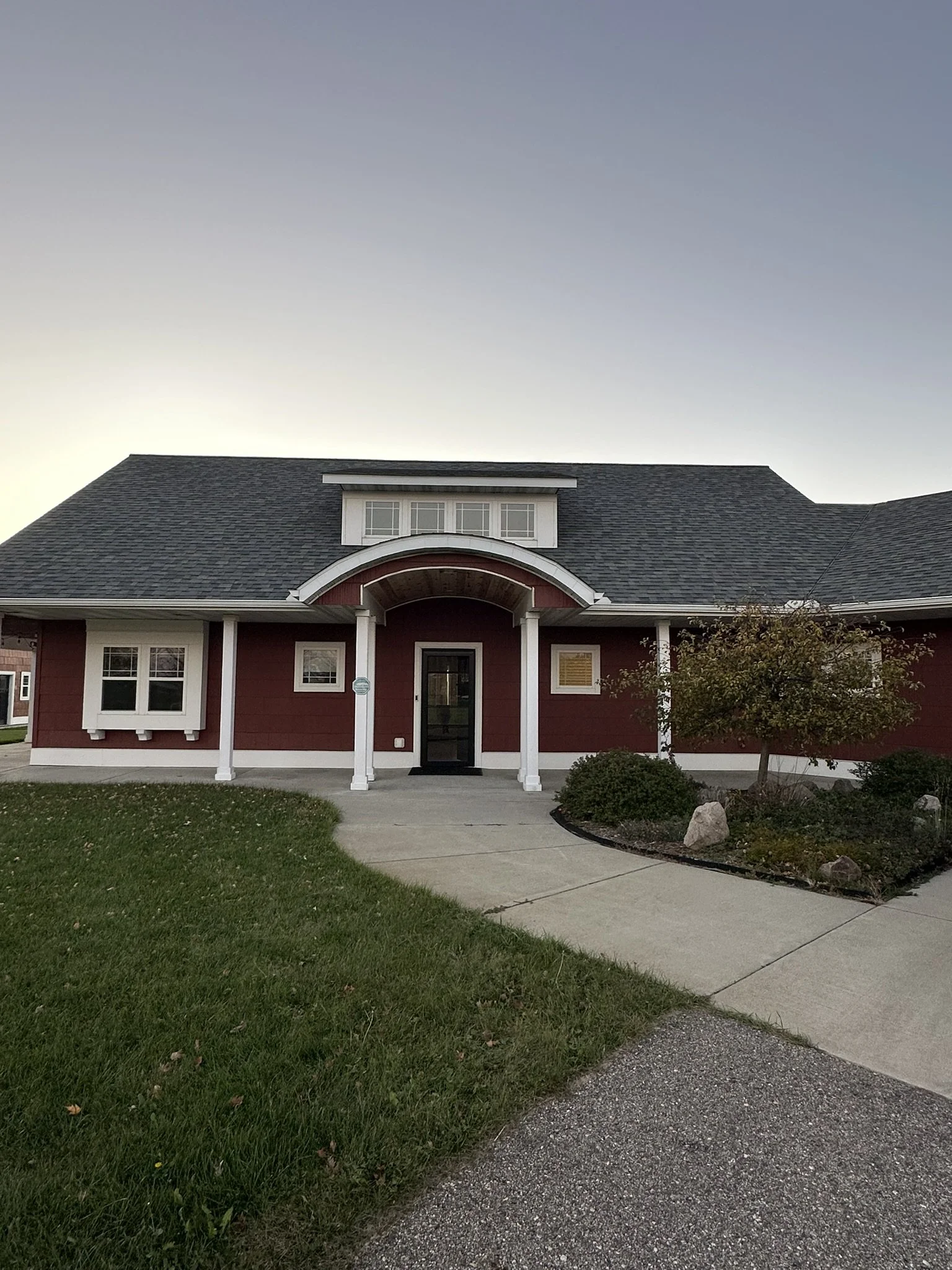 Front view of a red rental house with white trim, a black front door, a curved porch with white columns, a driveway, a small tree, and landscaped yard under a clear sky.