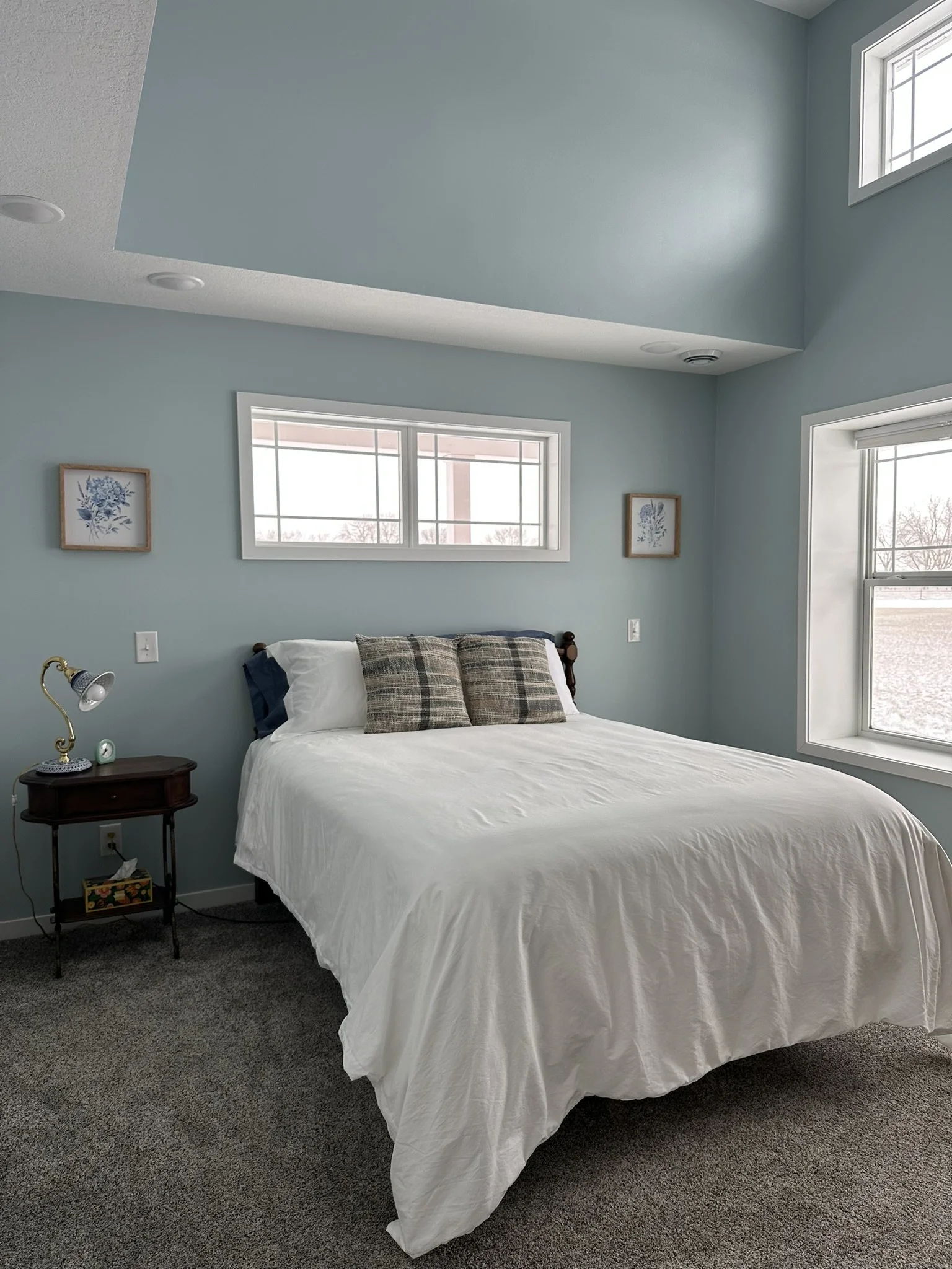A bedroom with light blue walls, a white bed with plaid pillows, and a small wooden nightstand with a lamp and small clock. There are two small framed art pieces on either side of a horizontal window above the bed, and a large window to the right. The room has gray carpet and a high ceiling.