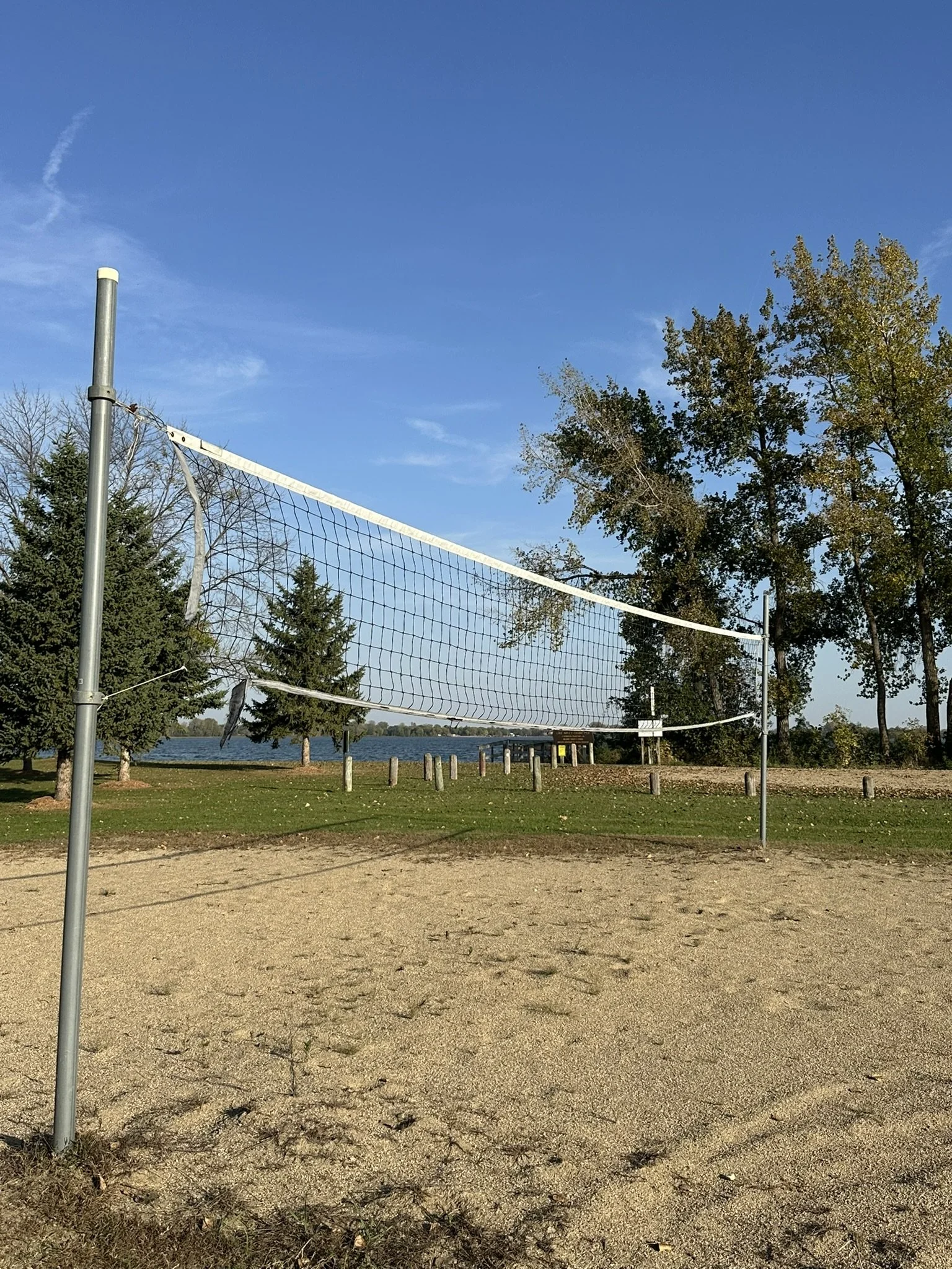 An outdoor sand volleyball court with a net, grass, trees, and a body of water in the background, under a blue sky.