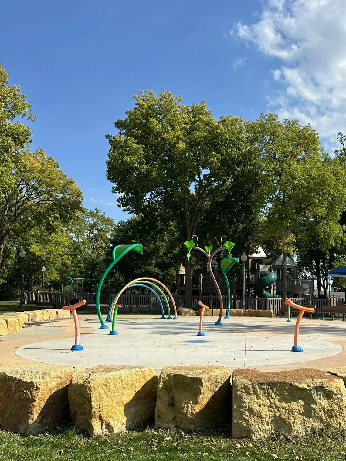 Children's splash pad playground with colorful water sources, trees in background, blue sky, and a slide in the distance.