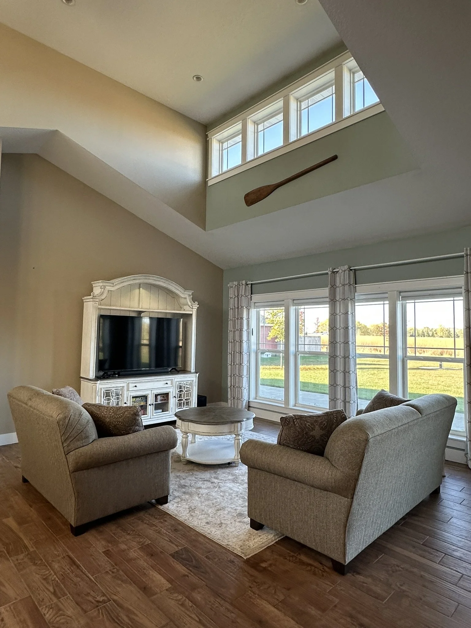 Living room with tall ceilings, two beige sofas, a white TV stand with a flat-screen TV, a round coffee table, floor to ceiling windows with curtains, and a view of a yard outside. An oar is mounted high on the wall near the ceiling.