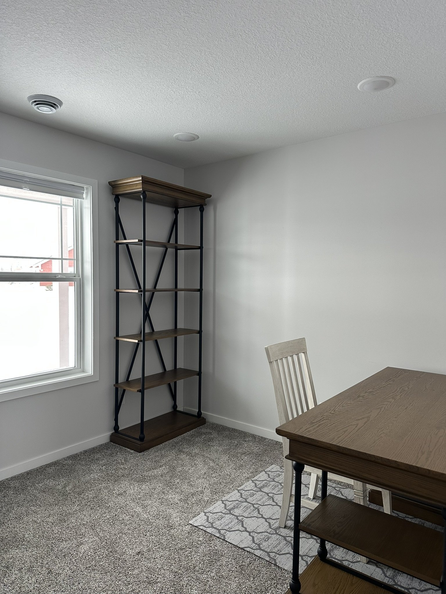 Empty room with a window, a tall bookshelf with wooden shelves and black metal frame, a table with a wooden top, a wooden chair, and a small rug on carpeted floor.