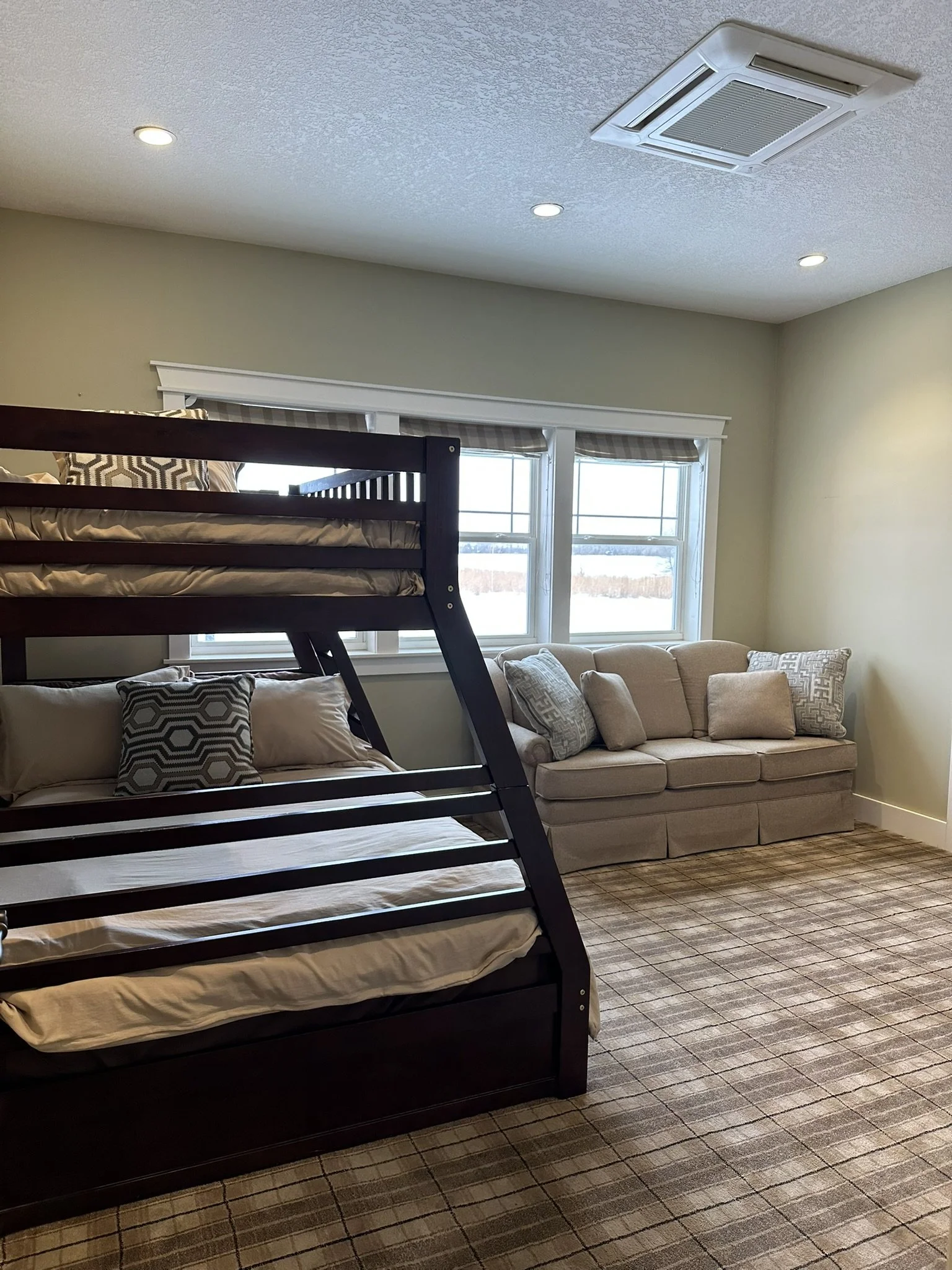 A room with a bunk bed over a couch and a windowed wall, a beige couch with pillows, and a carpeted floor. Office desk and mounted tv across from the couch.