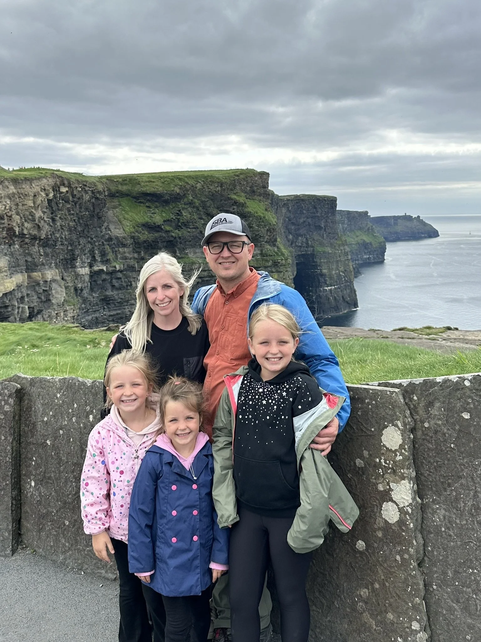 A family of six posing together outdoors at the Cliffs of Moher, Ireland, with green cliffs and the ocean in the background under a cloudy sky.
