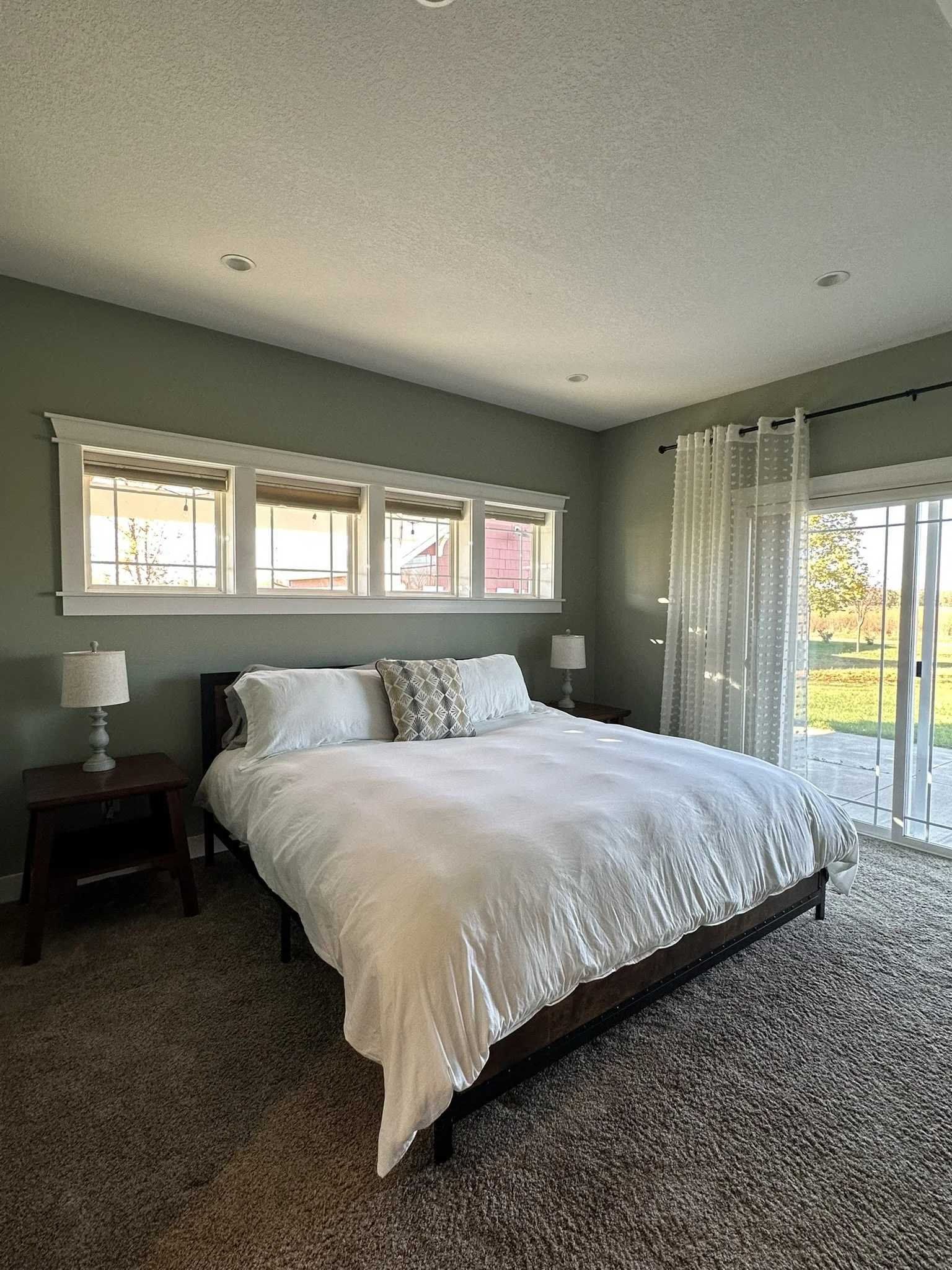 A bedroom with a bed, two side tables with lamps, green walls, windows, and sliding glass door with sheer curtains, overlooking a yard with trees.