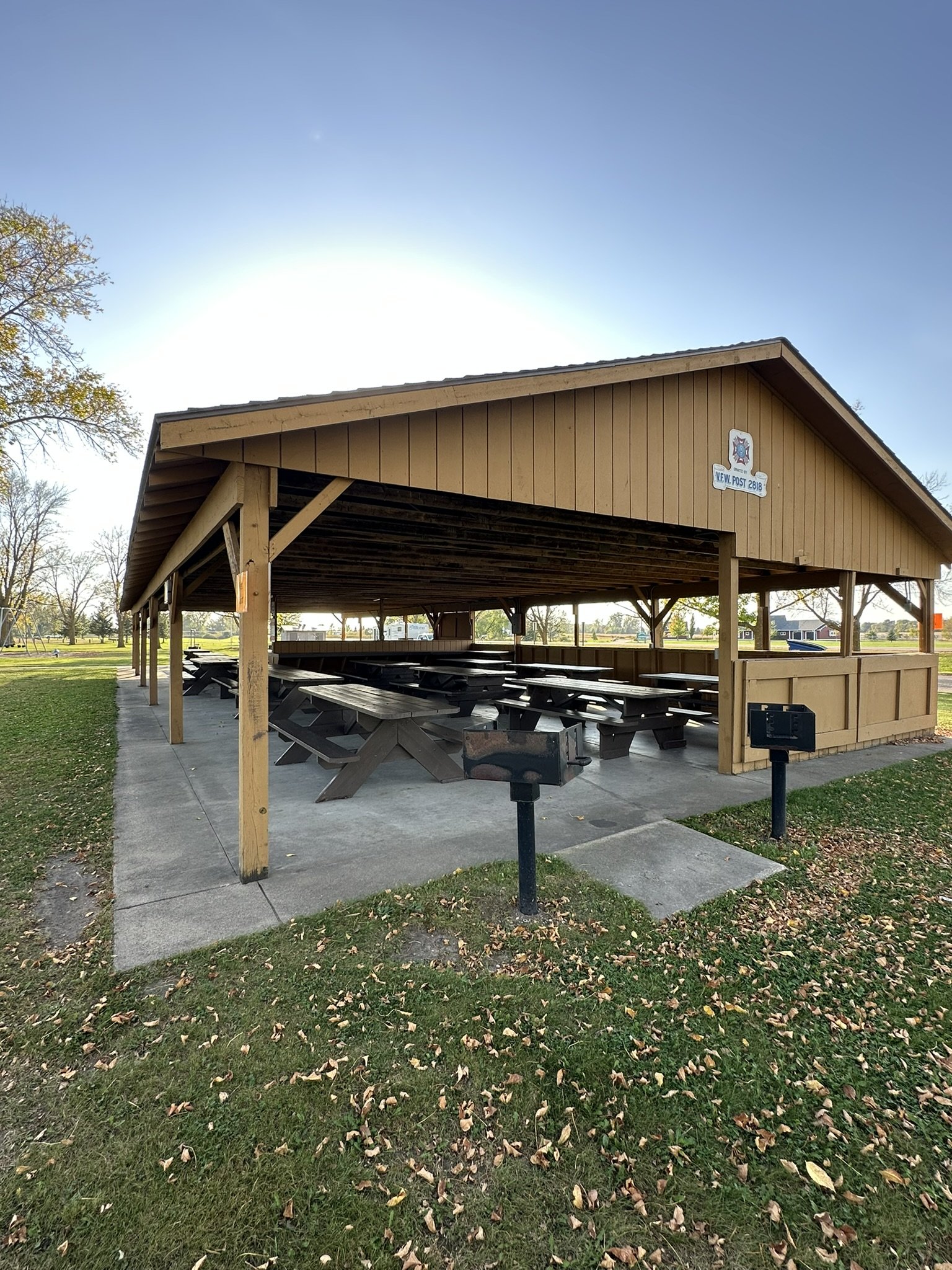 A wooden pavilion with picnic tables in a park, surrounded by grass and trees, with a bright blue sky and sun overhead.