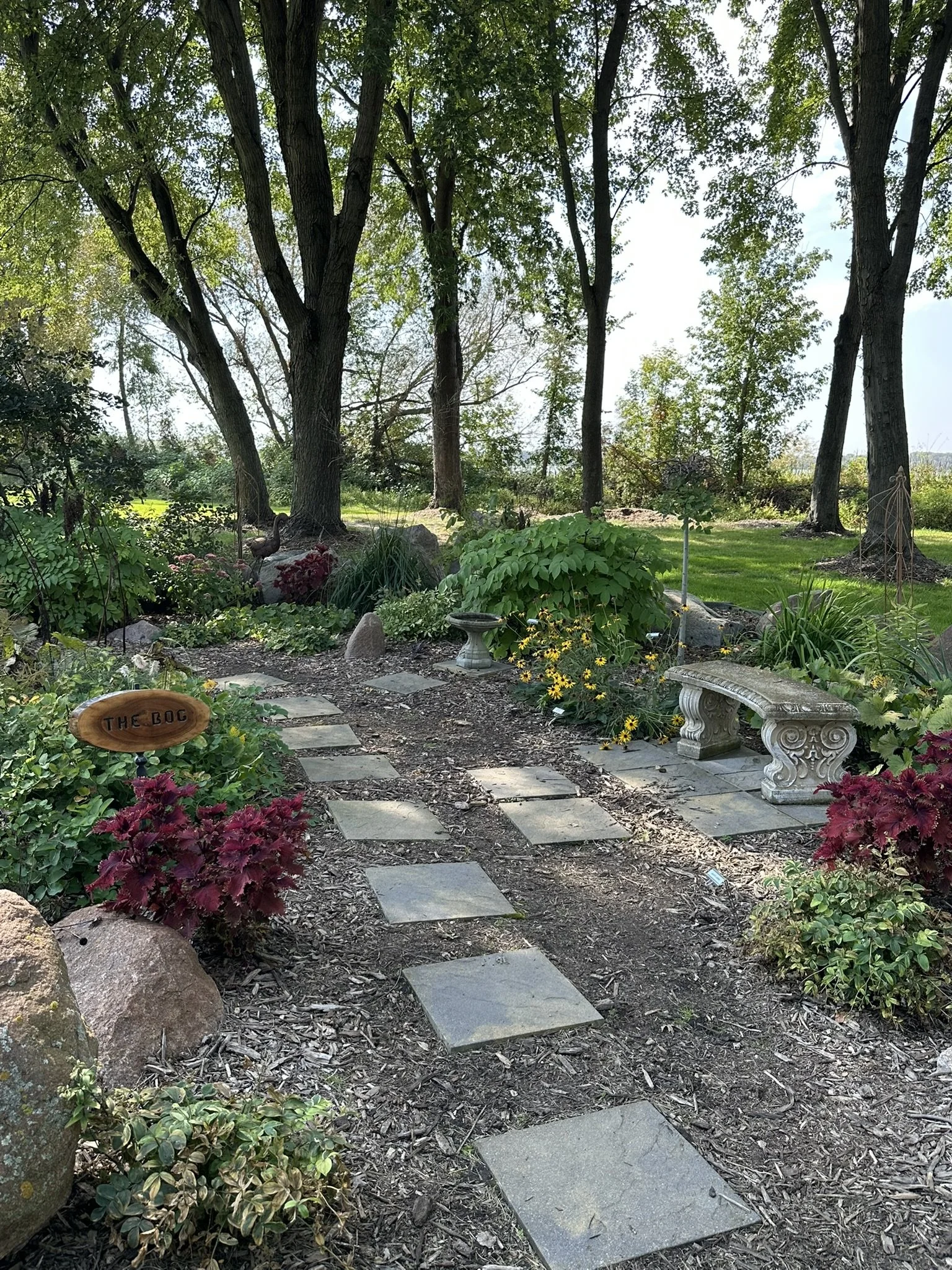 A garden pathway with stone pavers leading through lush greenery, pink and yellow flowers, and decorative benches under large trees.