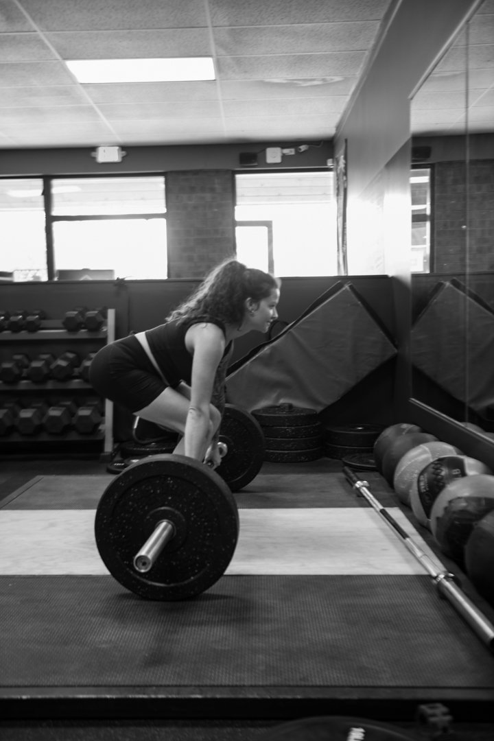 A woman is lifting a barbell in a gym with weight plates, surrounded by weights and gym equipment.