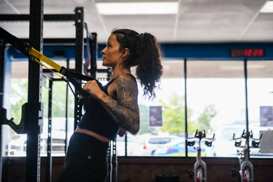 A woman with tattoos working out with suspension training straps inside a gym.