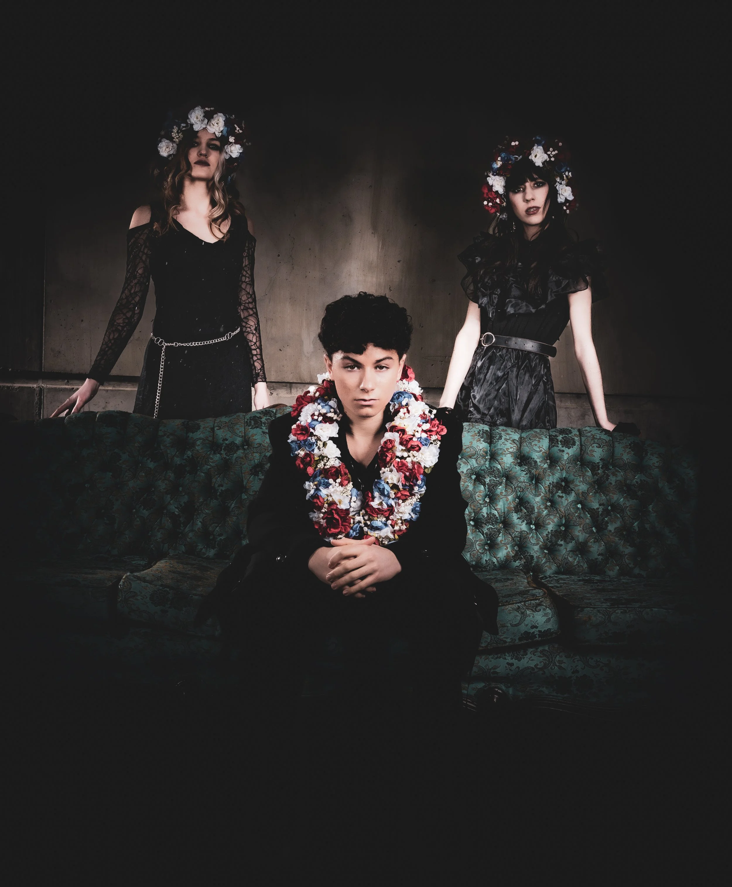 The band in dark attire and floral headpieces pose in a dimly lit room, with Jonah sitting on a vintage sofa in the foreground while Quinn and Aria are standing behind him.
