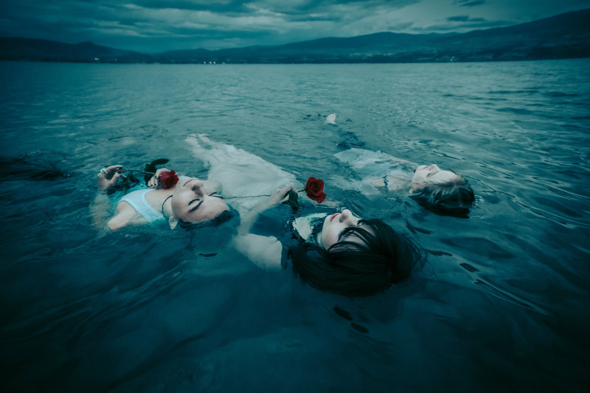 The band floating on a body of water, each holding a red rose, with mountains and cloudy sky in the background.