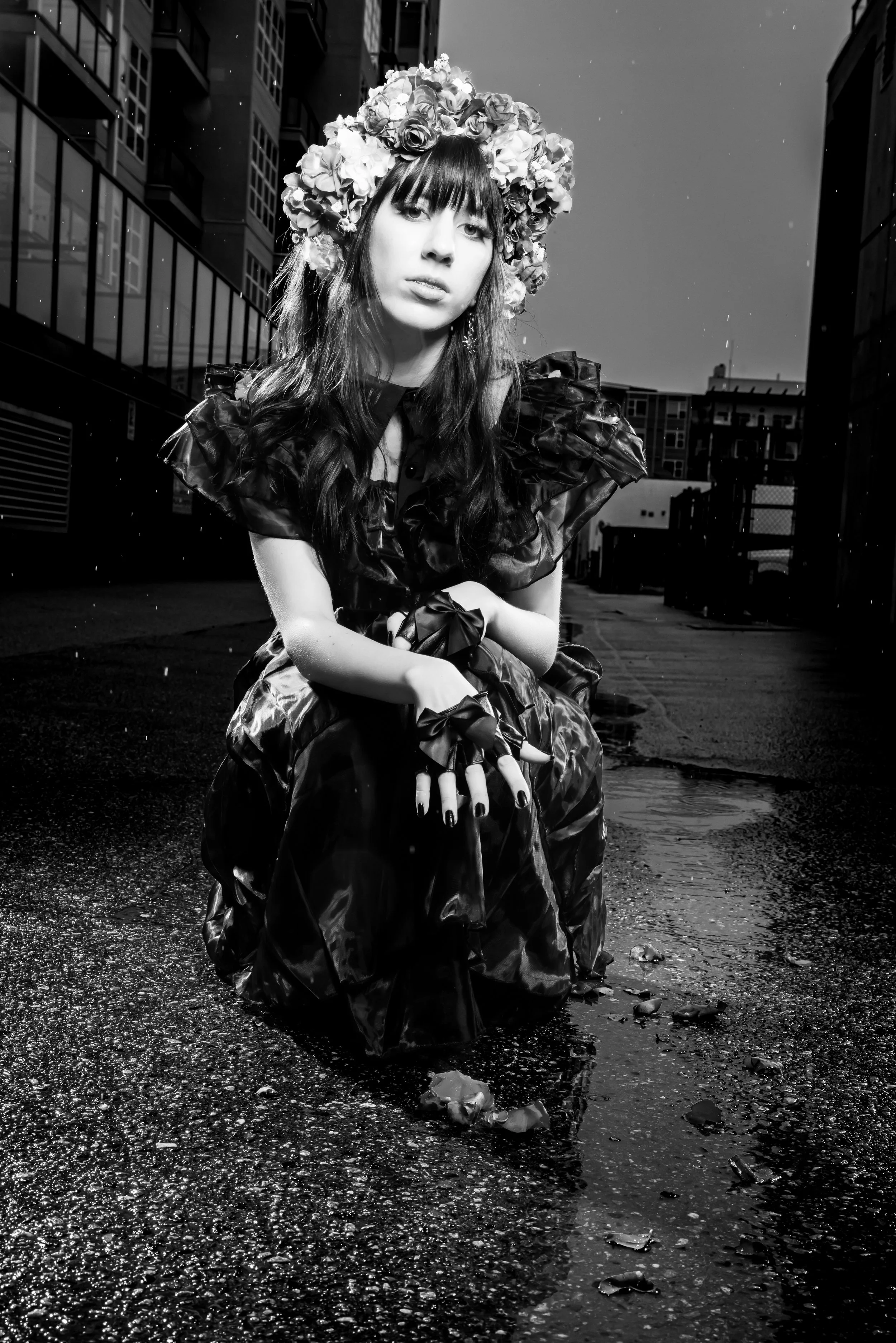 A woman with long dark hair and a floral crown kneeling on a wet city street at night, wearing a dark ruffled dress and matching gloves with ribbons.