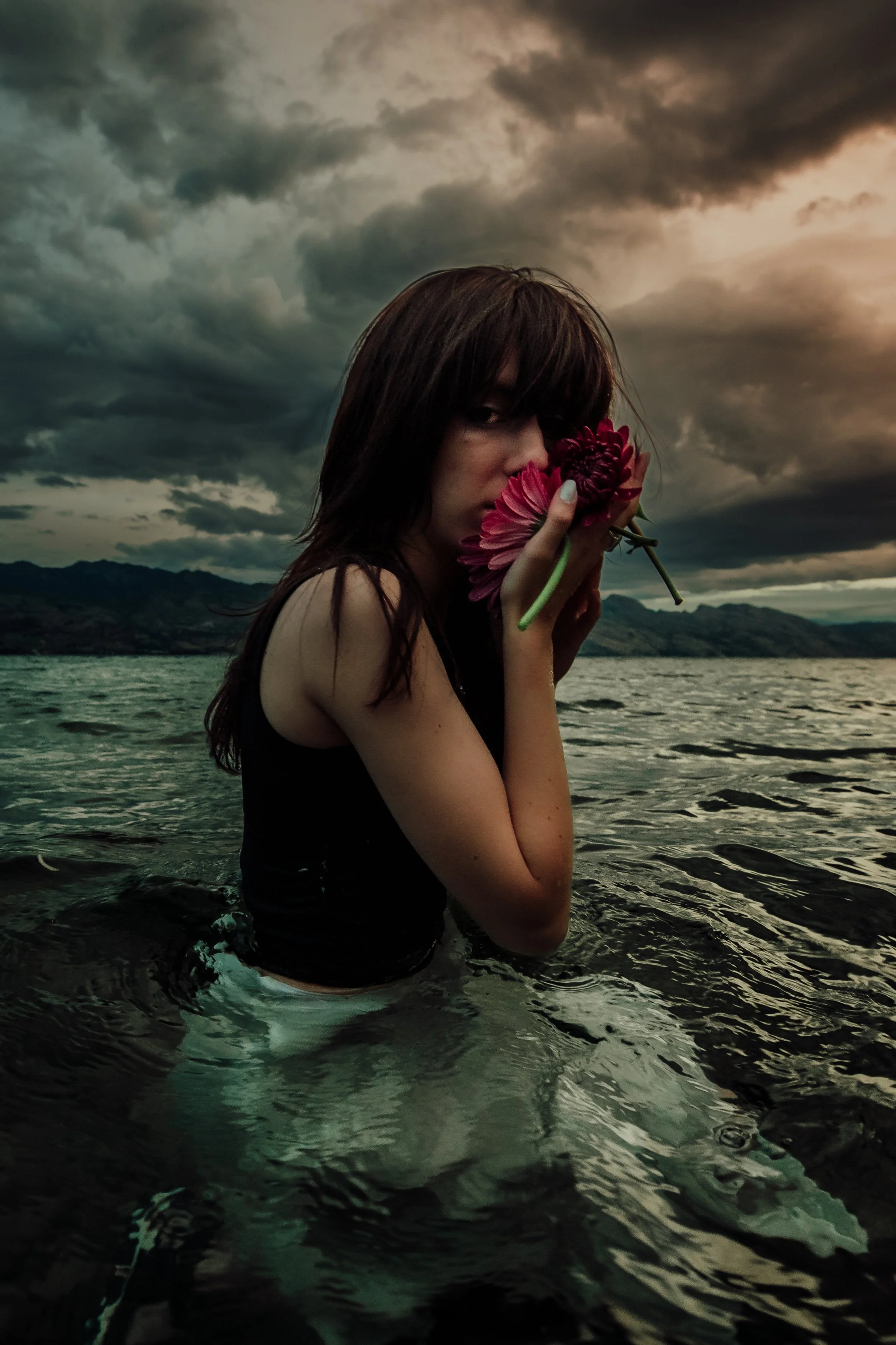 A woman with dark hair stands in a body of water at sunset, holding pink and purple flowers close to her face, with dark stormy clouds above and mountains in the distance.