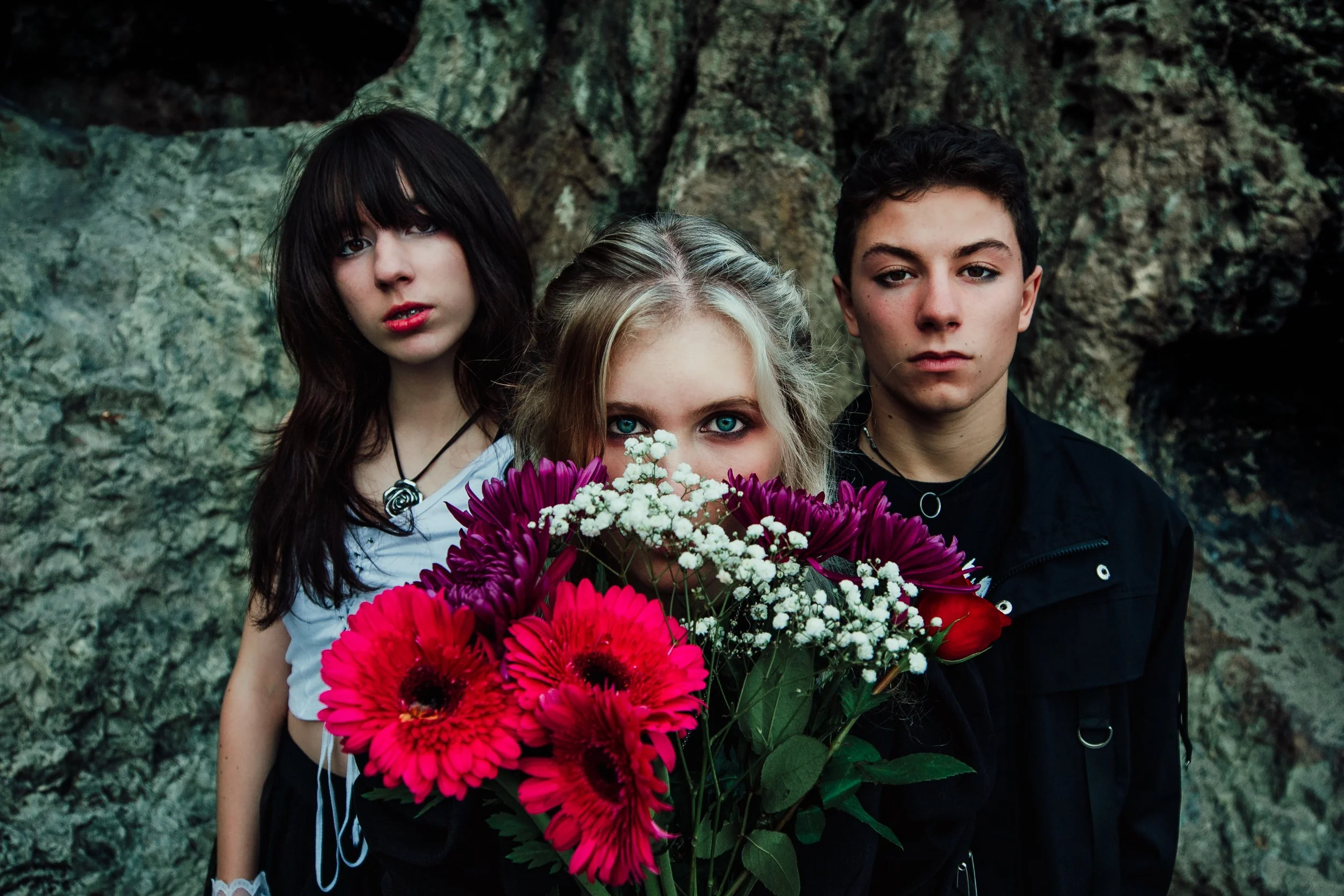 Three teenagers standing in front of a rocky background, with the girl in the middle holding a large bouquet of colorful flowers.