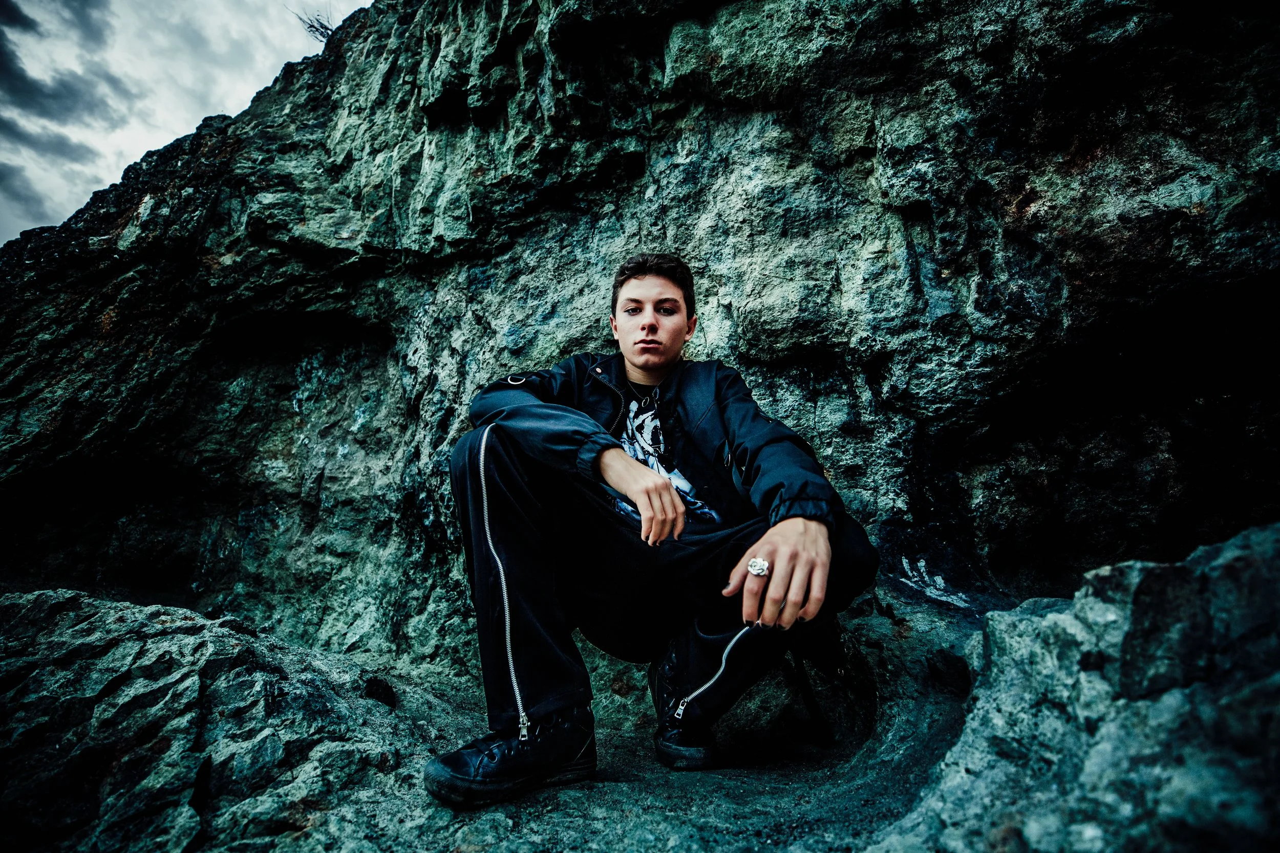 Young man wearing black clothing crouching on rocky terrain in front of a large rock wall, with cloudy sky overhead.