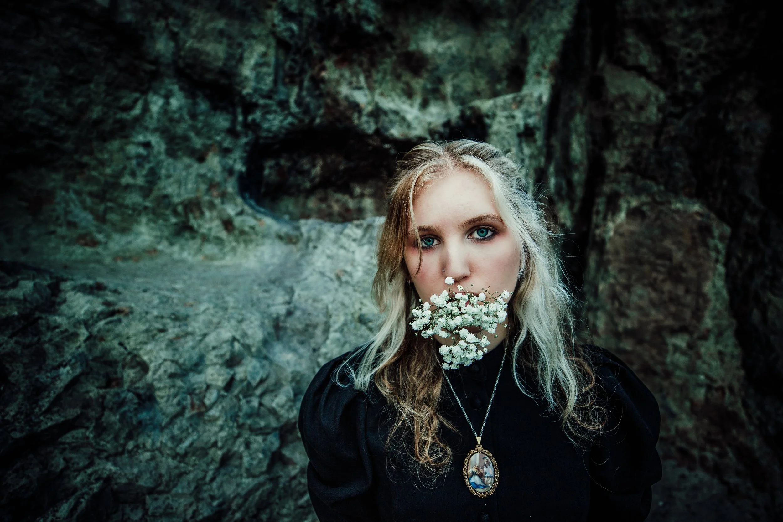 A young woman with blonde hair and blue eyes holding a cluster of white flowers in her mouth, standing in front of rocky cave entrance.