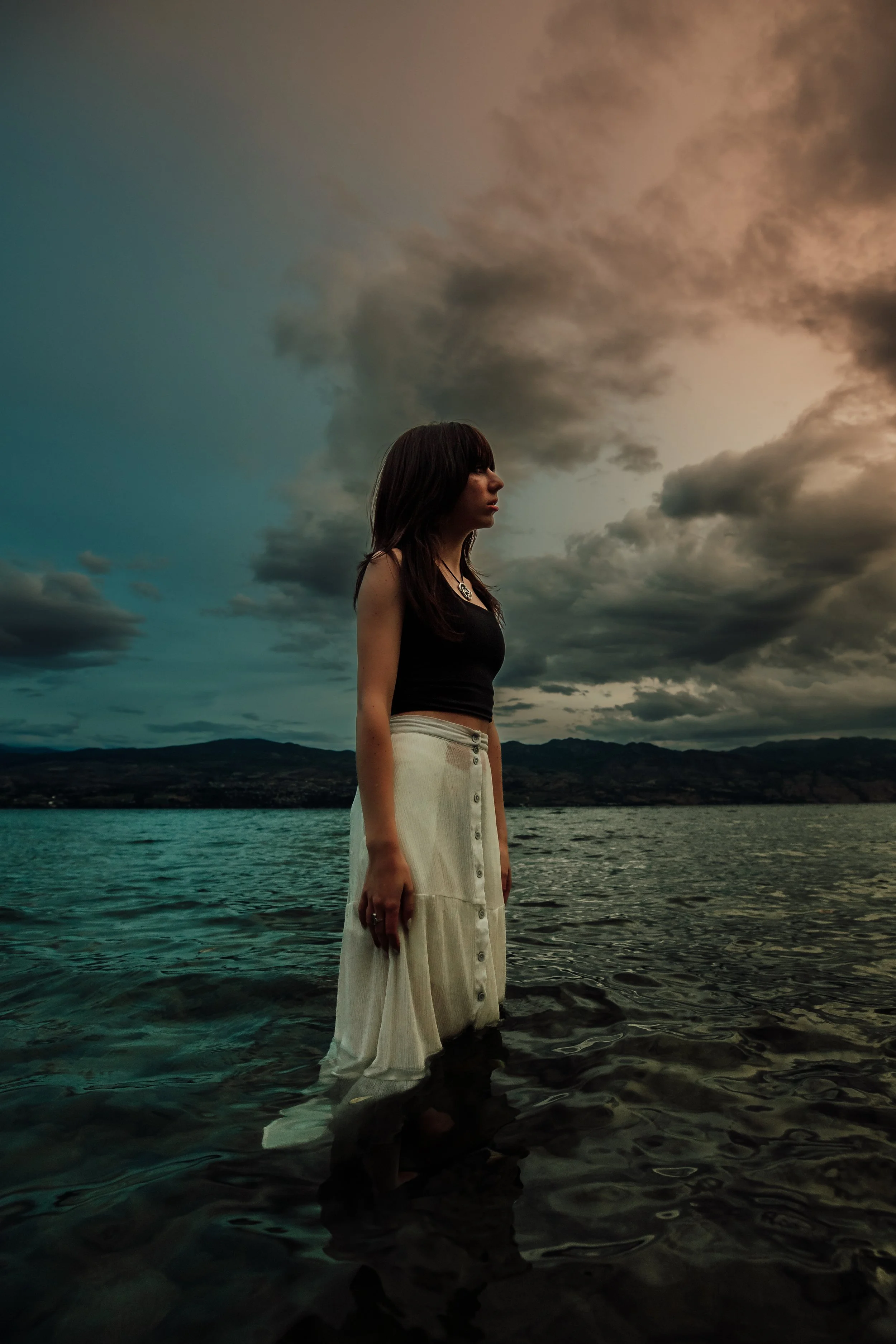 A woman standing in water at a lake during cloudy weather, wearing a black tank top and a long cream skirt, with dark hair and a serious expression.