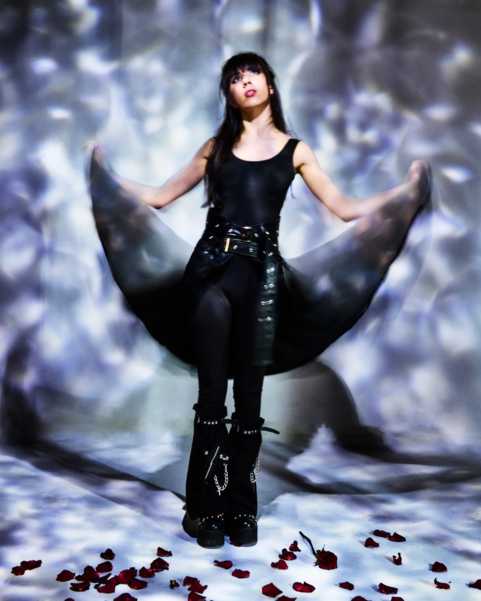 A woman with long dark hair wearing a black sleeveless top, leather pants, and platform boots is standing with her arms spread, holding a flowing black fabric. She is surrounded by falling red rose petals on the floor and a blurred, abstract backgrou