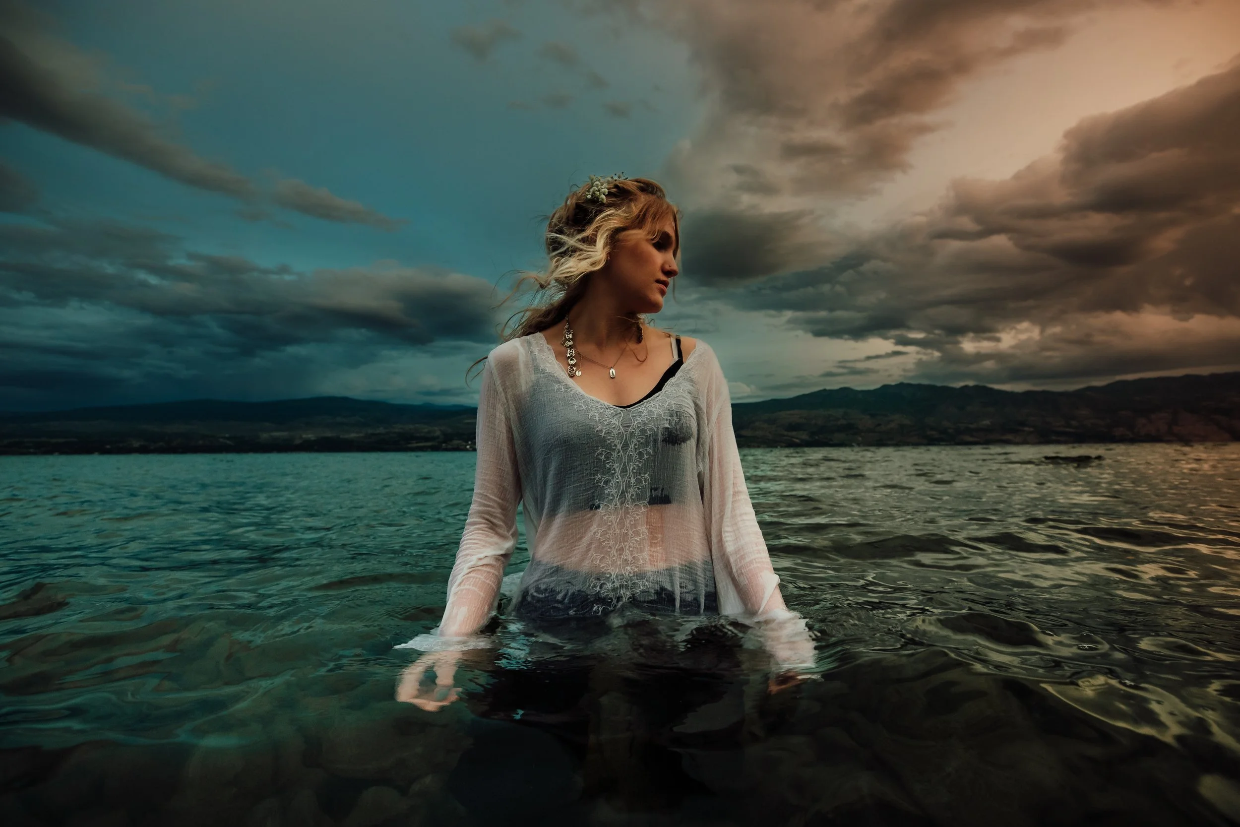 A woman in a white sheer top and black underneath stands in a body of water with a mountainous landscape and a dark, cloudy sky in the background.