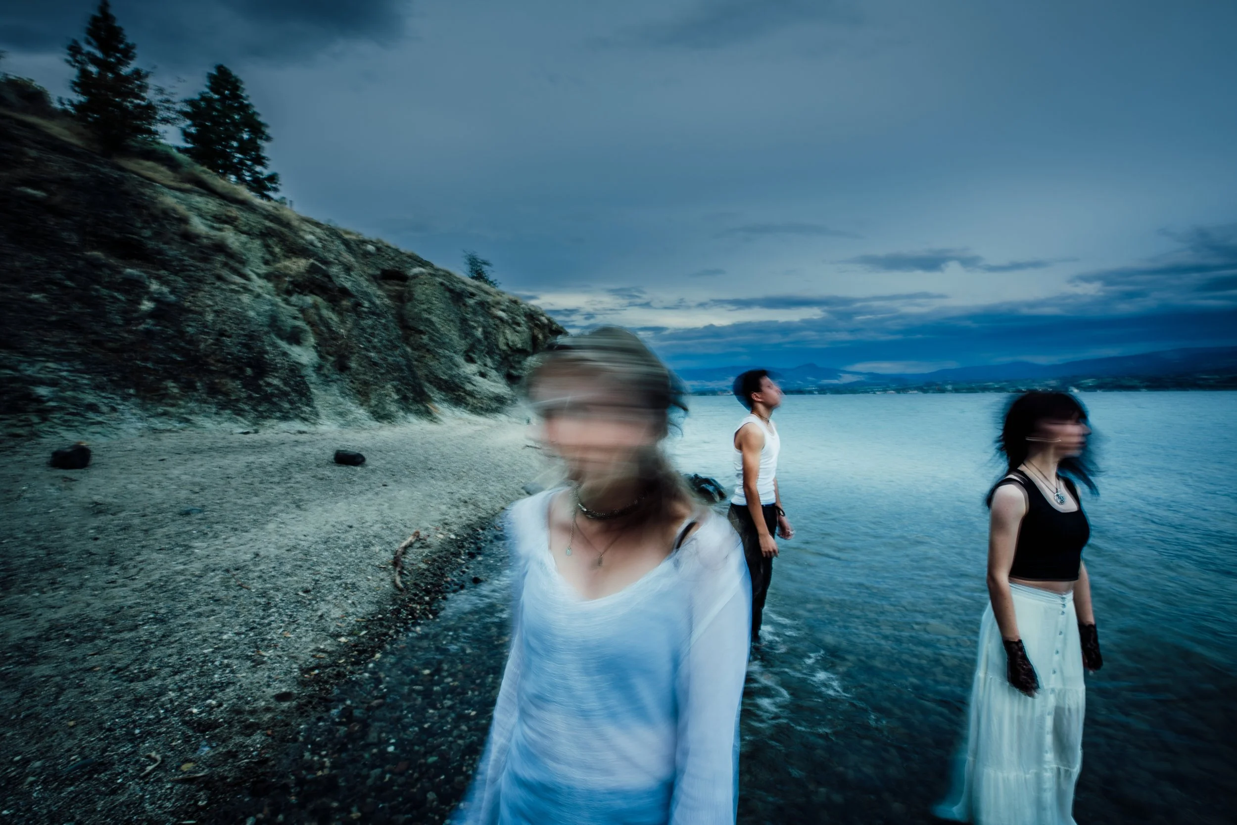 The band standing in shallow water at the beach during twilight, with a rocky cliff to their left and mountains in the distance across the water.