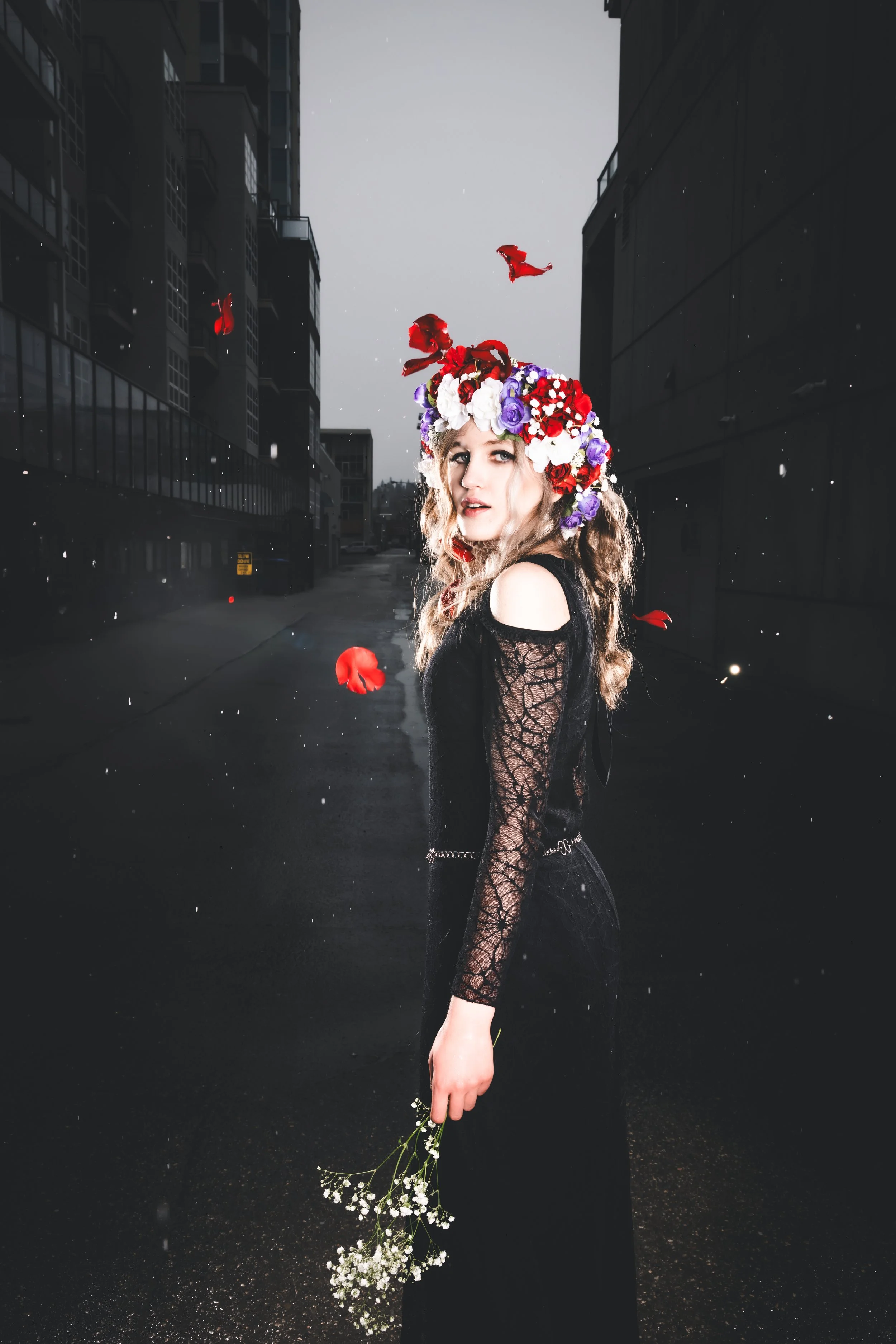 A woman standing in a dark, urban alleyway at dusk, wearing a black lace dress and a large, colorful flower crown, holding a small bouquet of white flowers.