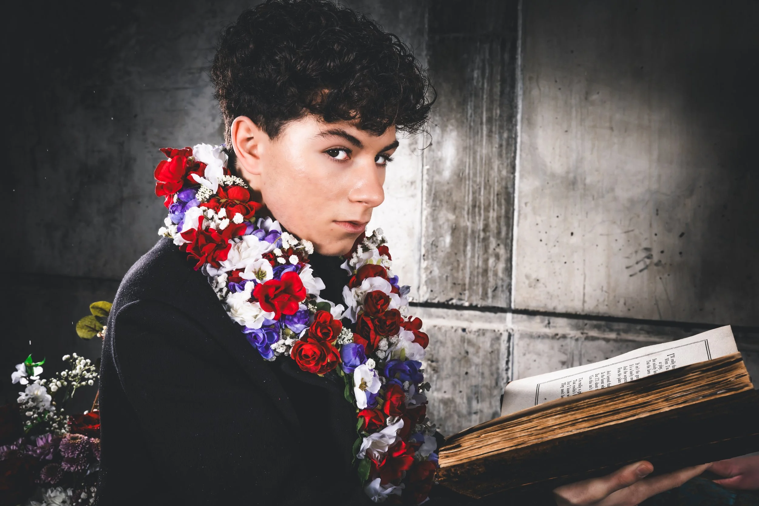 A person with dark curly hair wearing a black jacket and a colorful flower necklace, holding an old open book in front of a concrete wall.